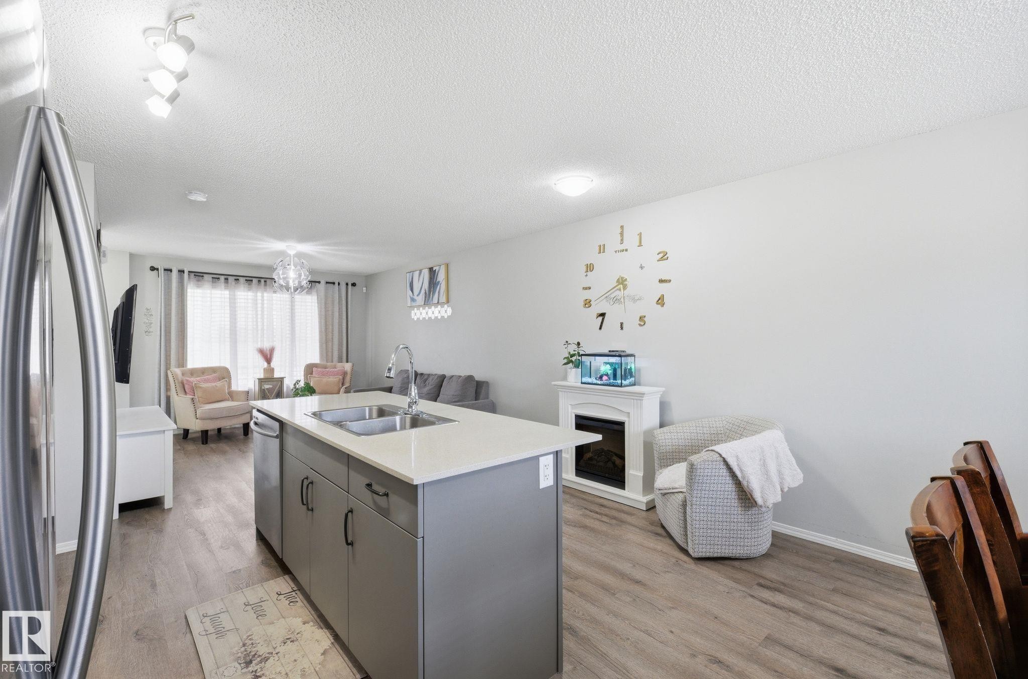 This open-concept area features a kitchen island with a double sink and light-colored countertop, alongside wood-look flooring throughout - 4636 175 Ave, Edmonton, AB - Indoor Photo Showing Kitchen With Double Sink