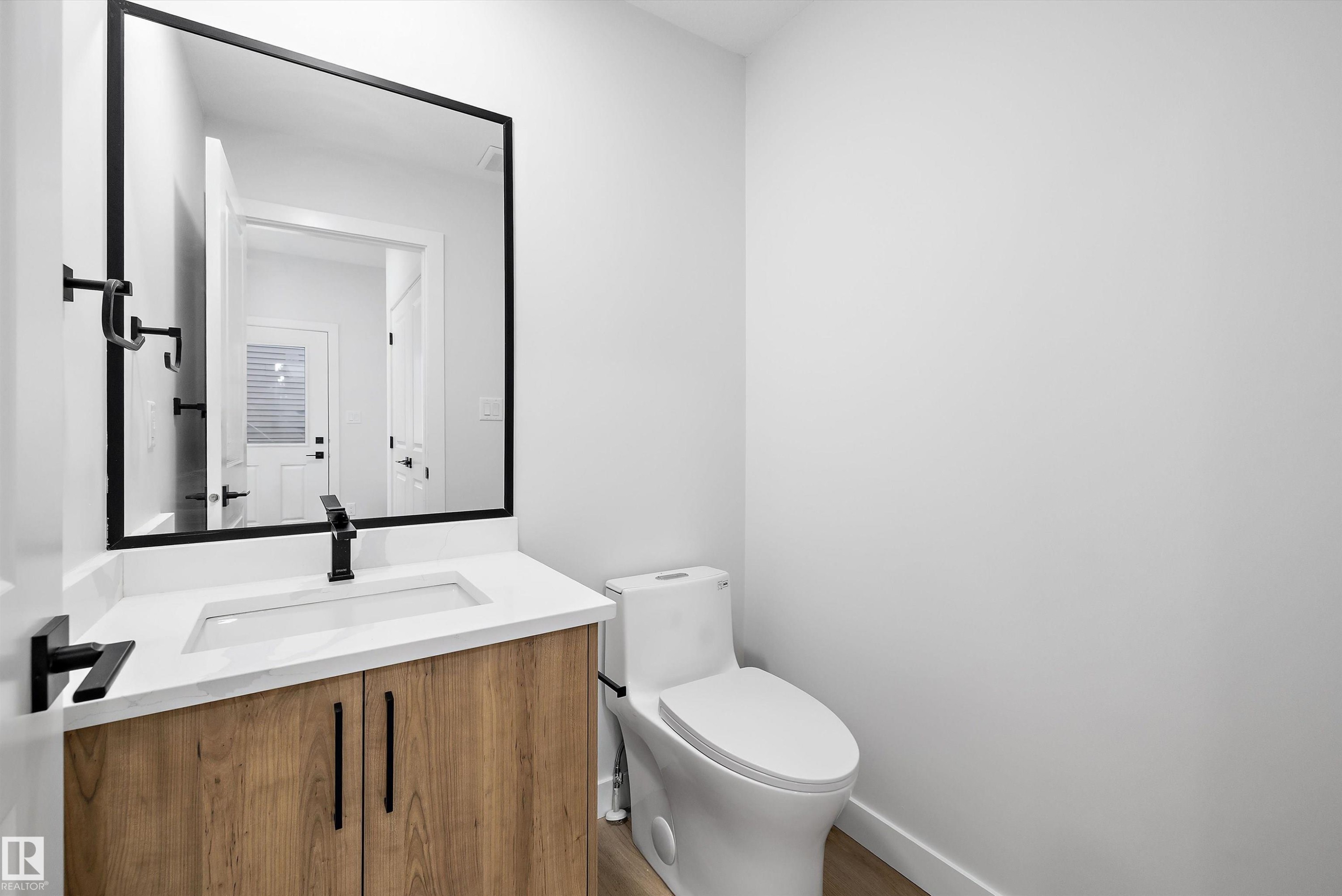 Powder room featuring a wood-finish vanity with a white countertop and integrated sink, a large black-framed mirror, and a modern toilet - 954 Elderberry Landing, Edmonton, AB - Indoor Photo Showing Bathroom