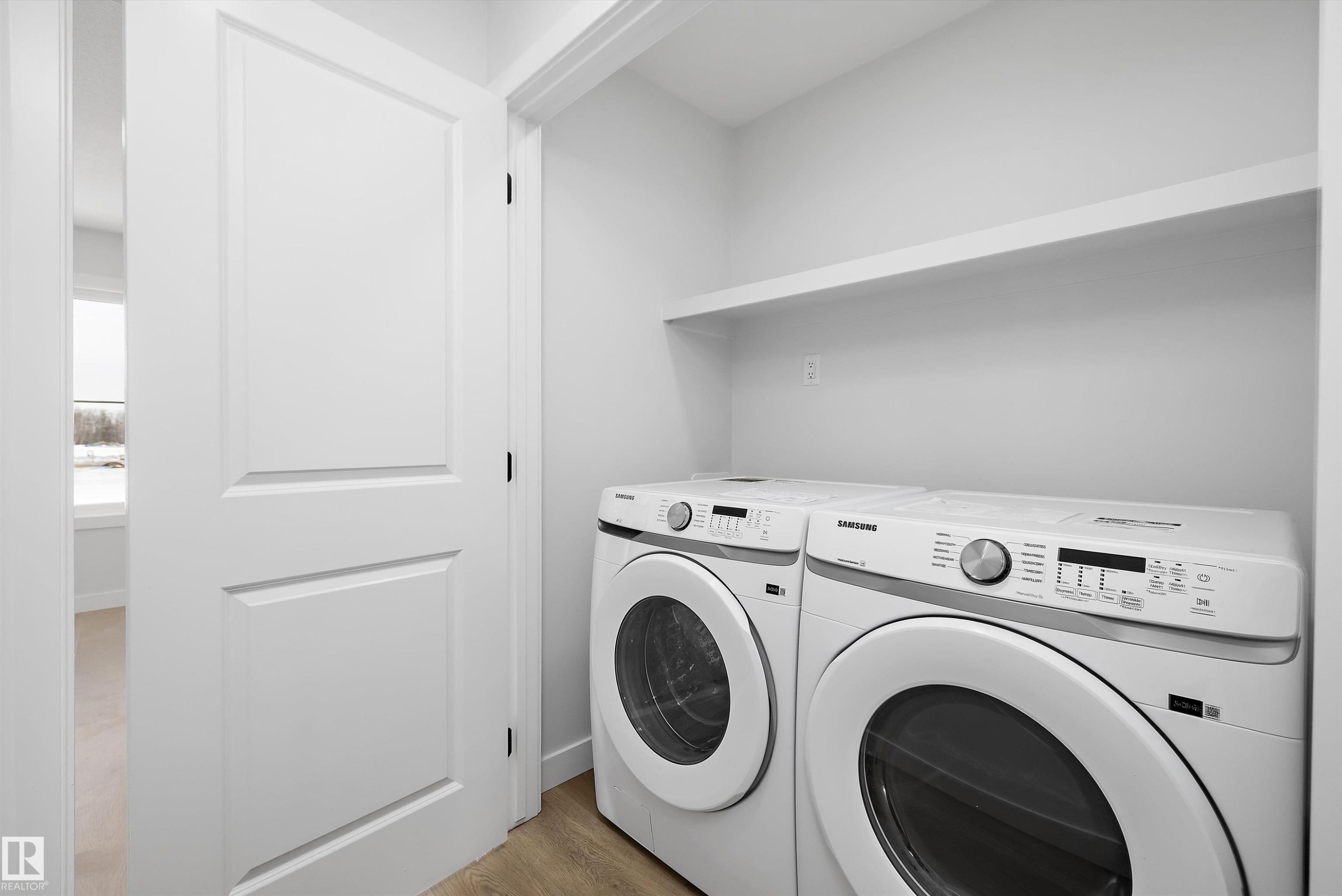 Dedicated laundry area featuring a front-loading washer and dryer, integrated shelving, and wood-finish flooring - 954 Elderberry Landing, Edmonton, AB - Indoor Photo Showing Laundry Room