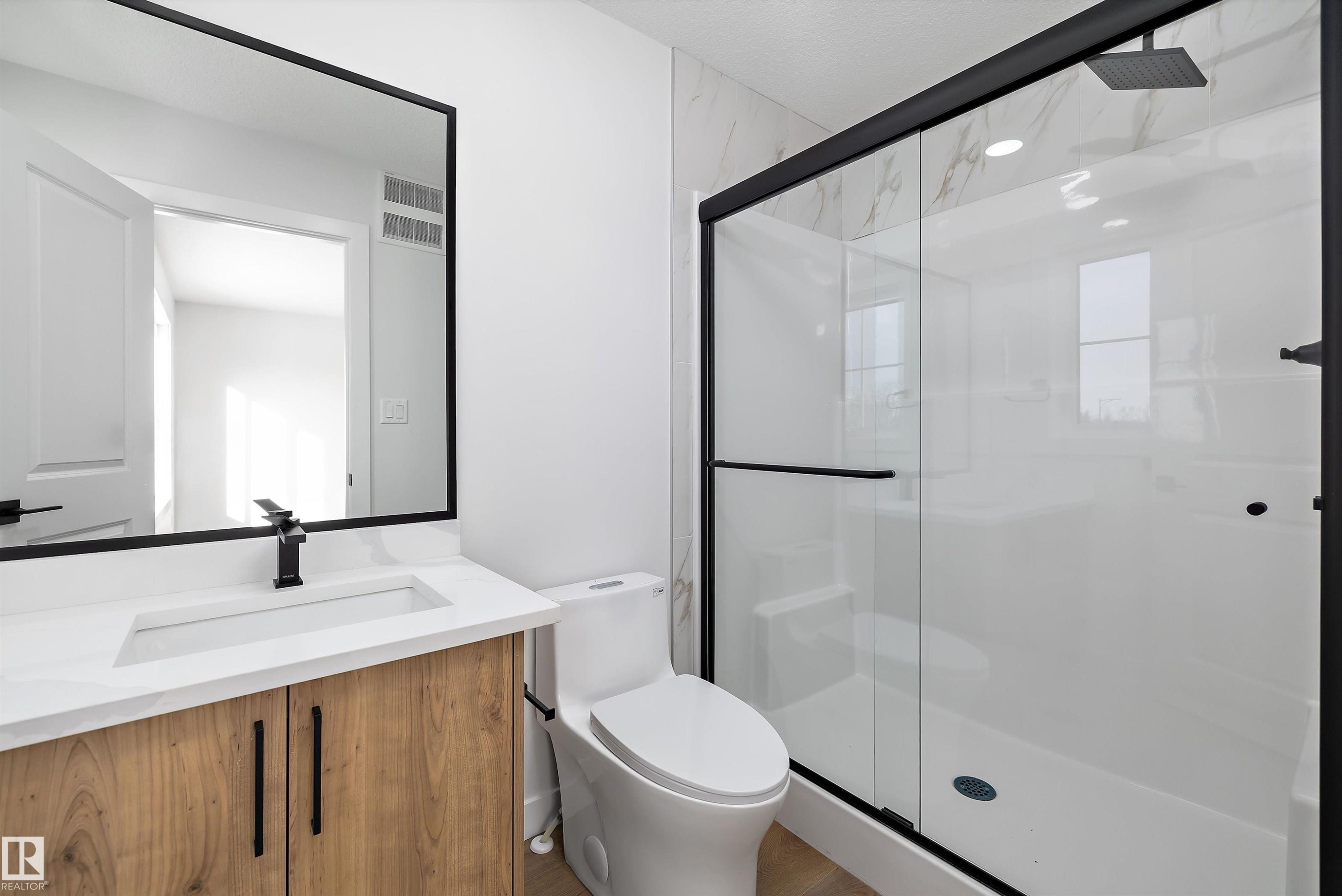 Contemporary bathroom featuring a wood-finish vanity with a white countertop and integrated sink, a large framed mirror, and matte black fixtures - 954 Elderberry Landing, Edmonton, AB - Indoor Photo Showing Bathroom