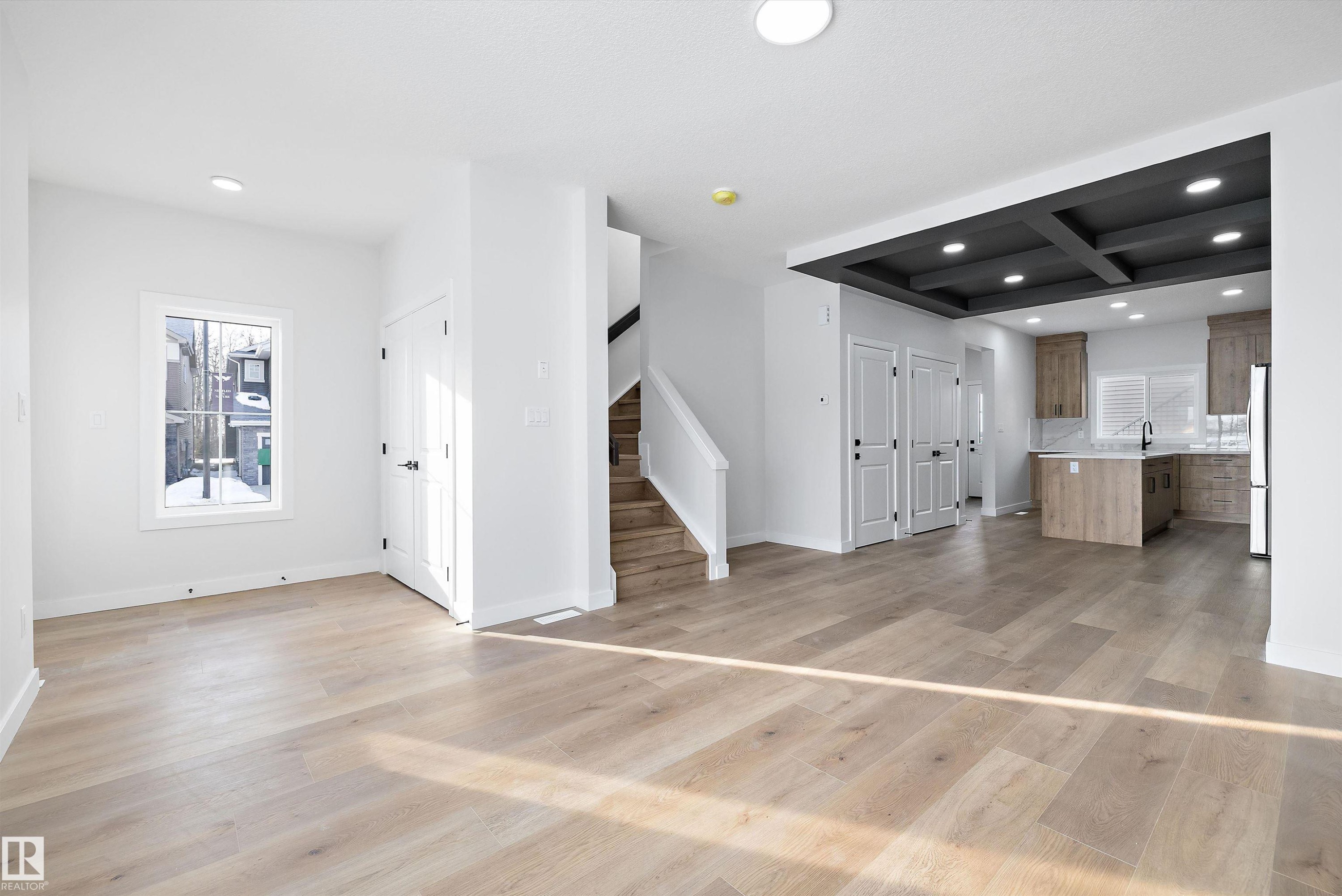 Open-concept main level featuring light wood-finish flooring, white trim, and a contemporary kitchen with wood-finish cabinetry, a central island, and a dark coffered ceiling - 954 Elderberry Landing, Edmonton, AB - Indoor Photo Showing Other Room