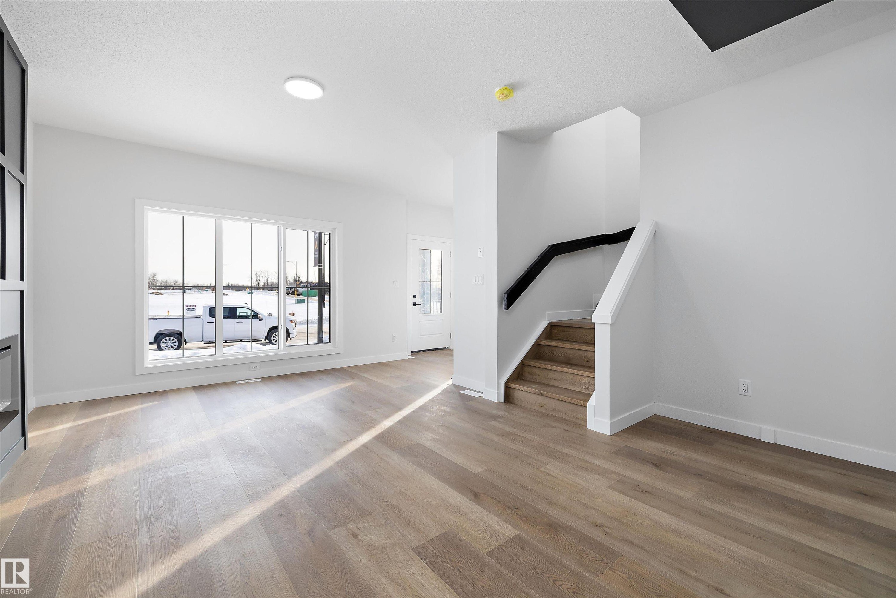 Open-concept living space featuring wood-finish flooring, a large multi-pane window, and a modern staircase with a black handrail - 954 Elderberry Landing, Edmonton, AB - Indoor