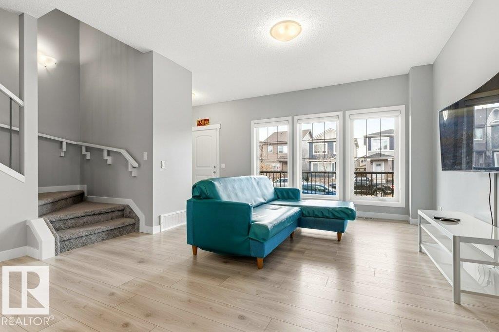 Bright living area featuring light wood-finish flooring, three large windows, a textured ceiling, and a carpeted staircase with a white railing - 17451 77 Street, Edmonton, AB - Indoor Photo Showing Living Room