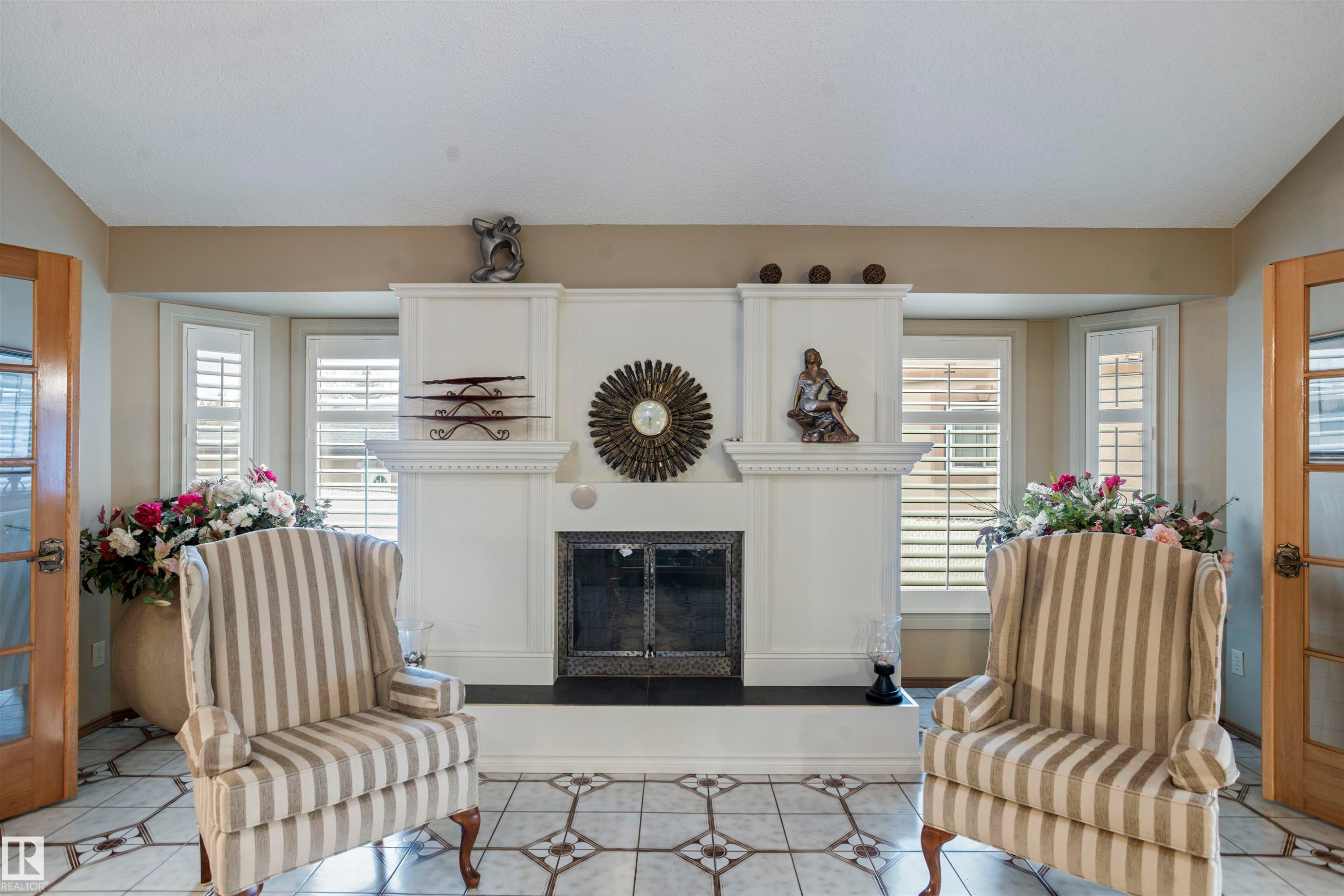 Ornate fireplace with a black hearth and decorative mantelpiece, flanked by two bay windows with plantation shutters - 9831 158 Avenue, Edmonton, AB - Indoor Photo Showing Living Room With Fireplace