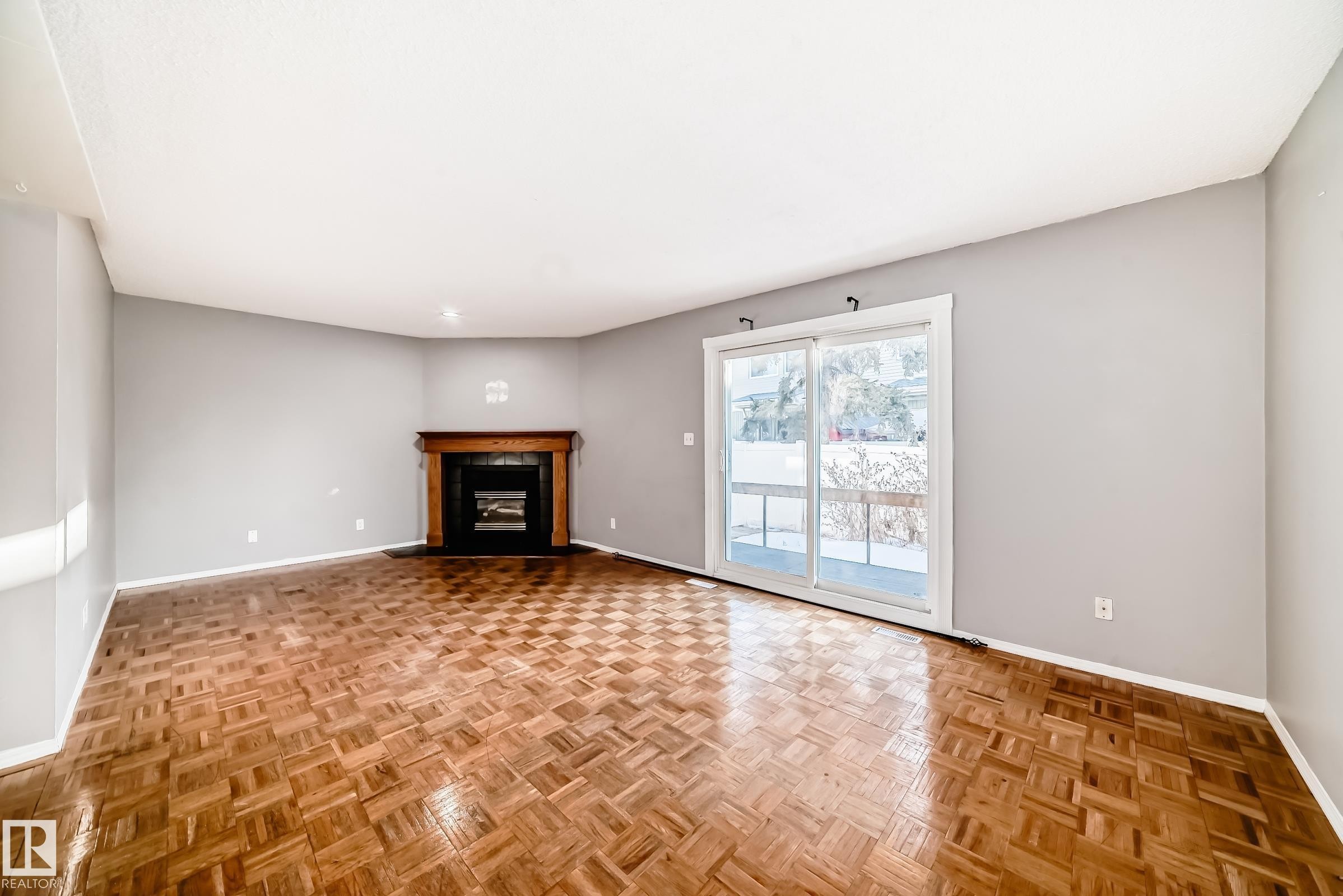 This bright room features parquet flooring, a corner fireplace with a dark wood mantel, and sliding glass doors that open to an exterior area - 8217 182 Street Nw, Edmonton, AB - Indoor With Fireplace