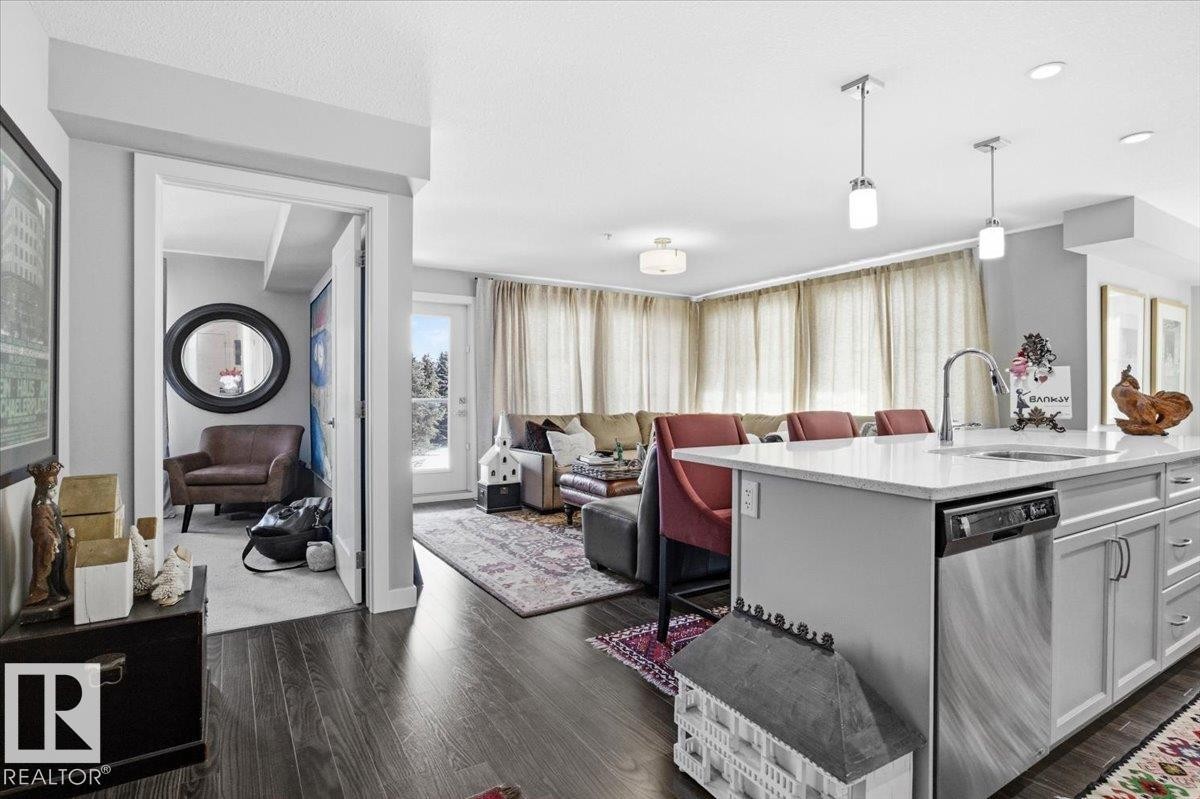 The property features hardwood floors, a kitchen island with a sink and pendant lighting, and a living area with large windows providing natural light - 211 5029 Edgemont Boulevard, Edmonton, AB - Indoor Photo Showing Kitchen With Upgraded Kitchen