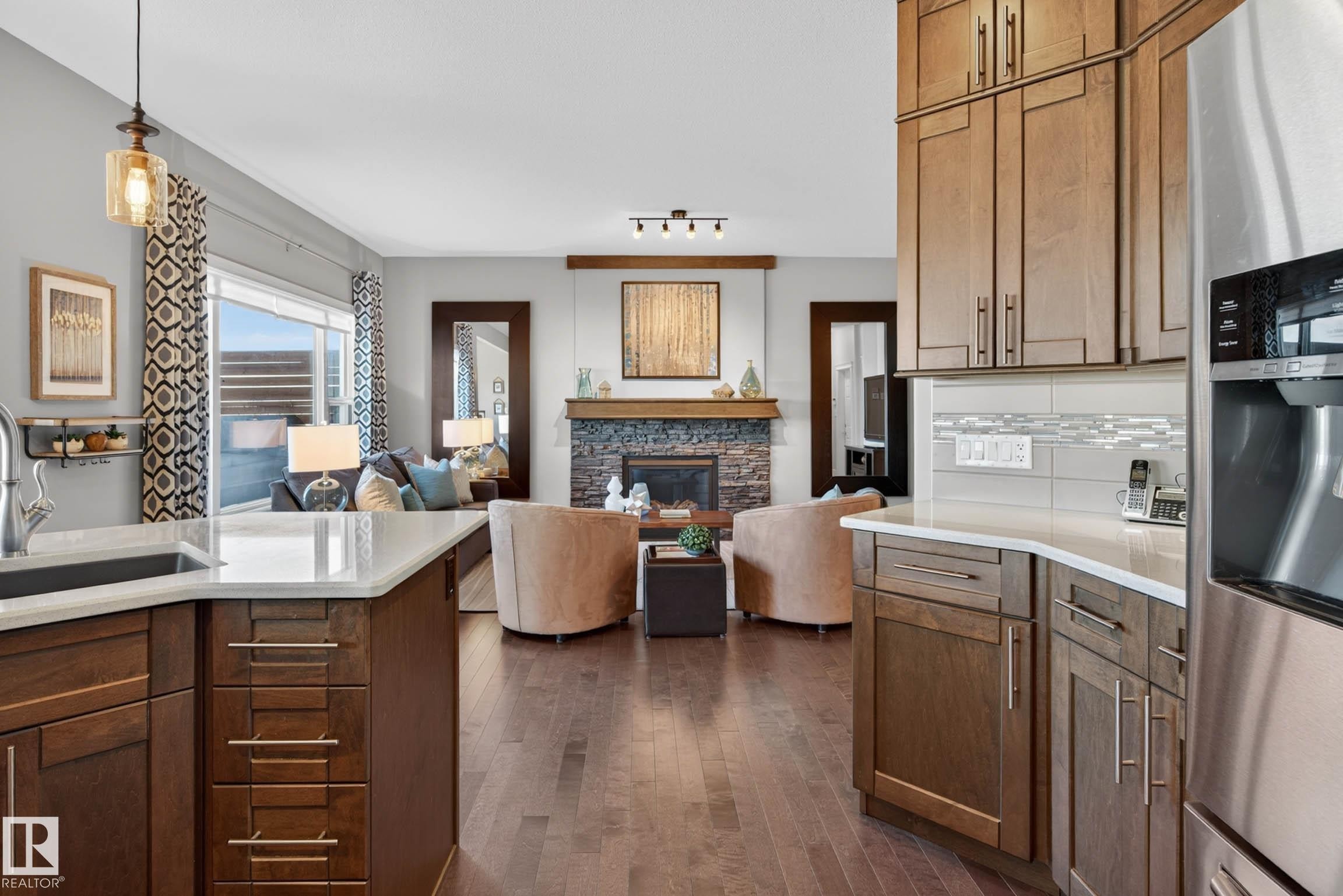 Open-concept living space featuring wood-finish flooring, a stone fireplace with a wood mantle, and a kitchen island with a built-in sink and white countertop - 2086 Redtail Common, Edmonton, AB - Indoor Photo Showing Kitchen