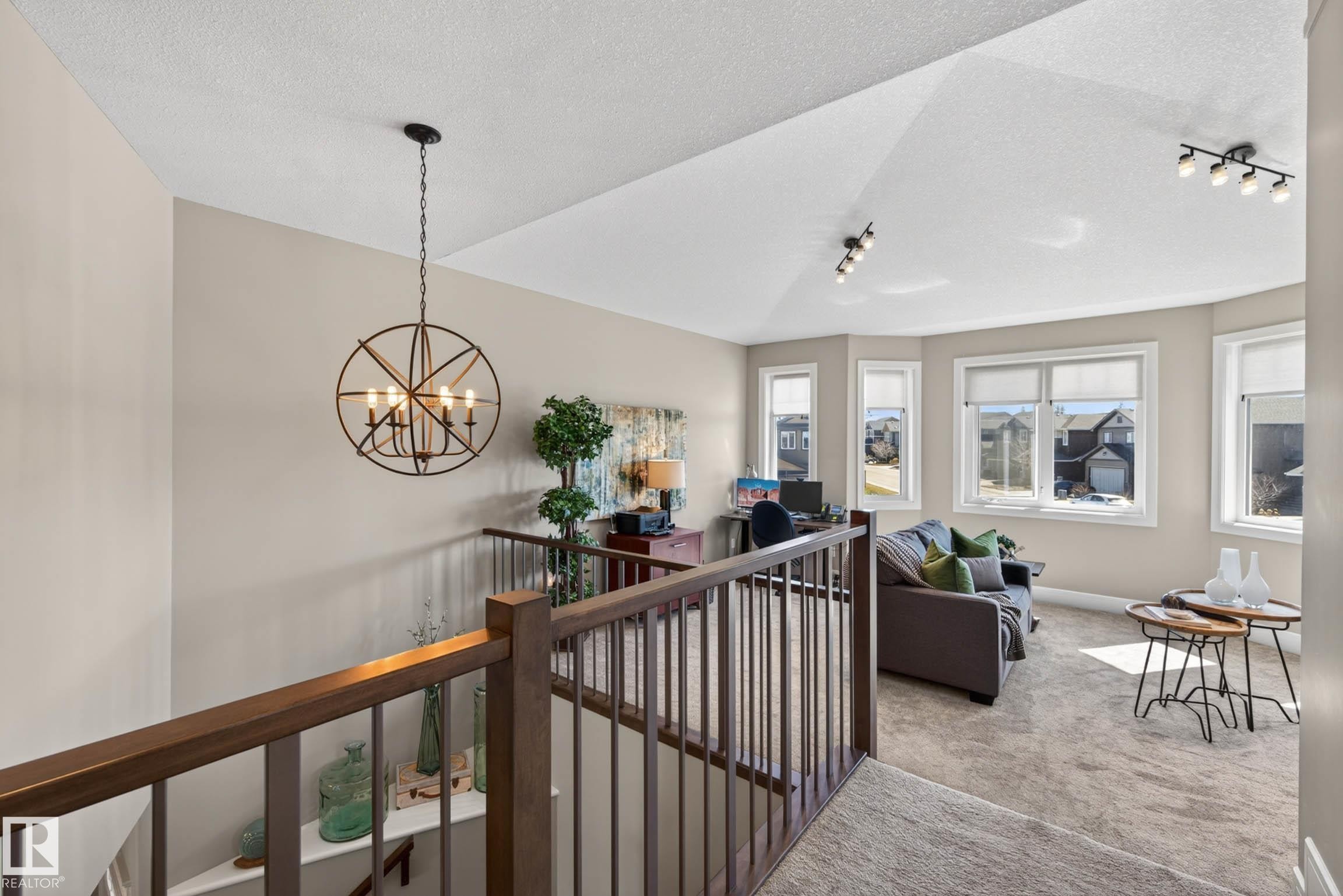 Open landing area featuring a modern chandelier, wood-finish railing, bay windows, textured carpet, and track lighting - 2086 Redtail Common, Edmonton, AB - Indoor Photo Showing Other Room