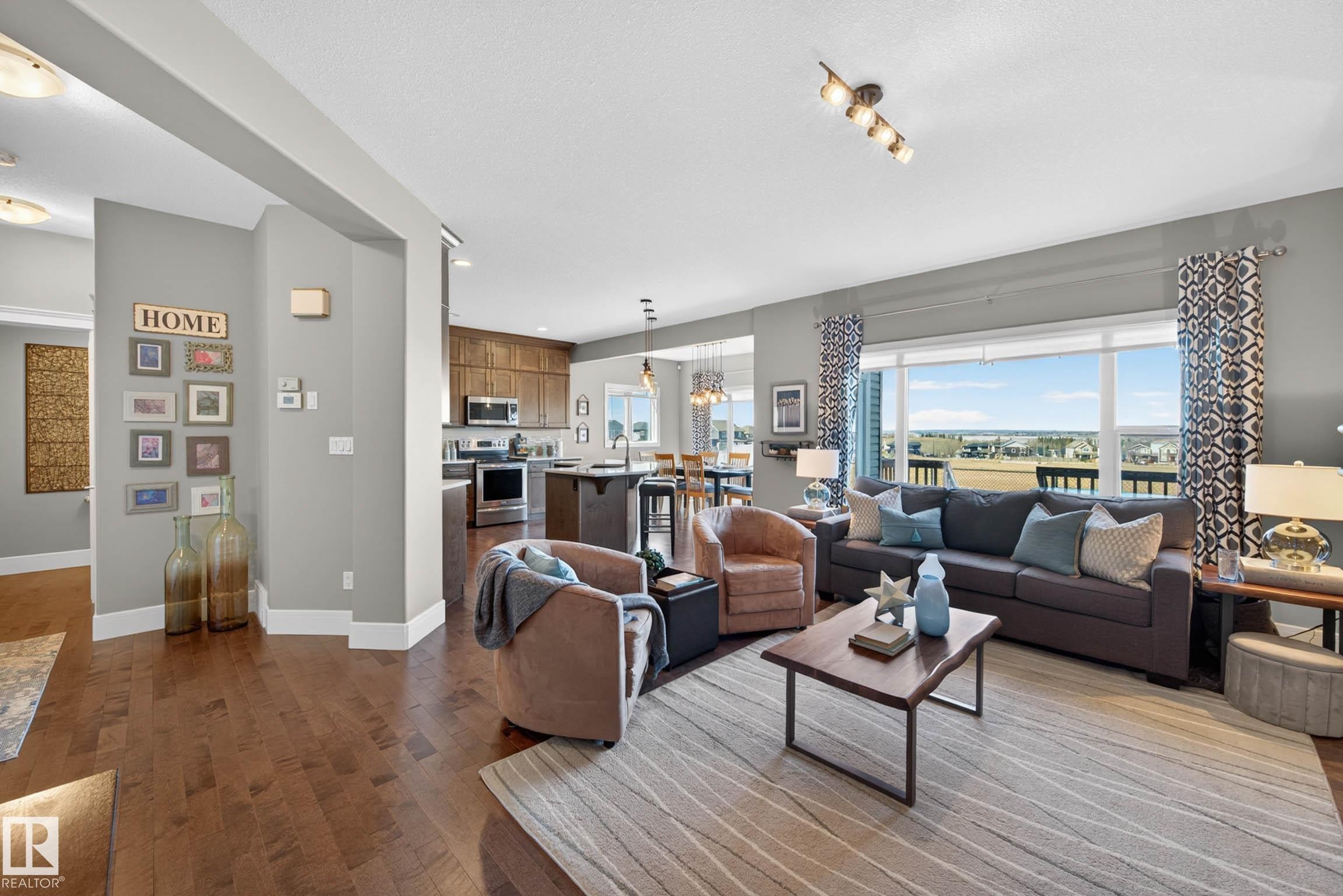 Spacious open-concept living area featuring wood-finish flooring, a large picture window, recessed lighting, and an expansive kitchen with built-in cabinetry and stainless steel appliances - 2086 Redtail Common, Edmonton, AB - Indoor Photo Showing Living Room
