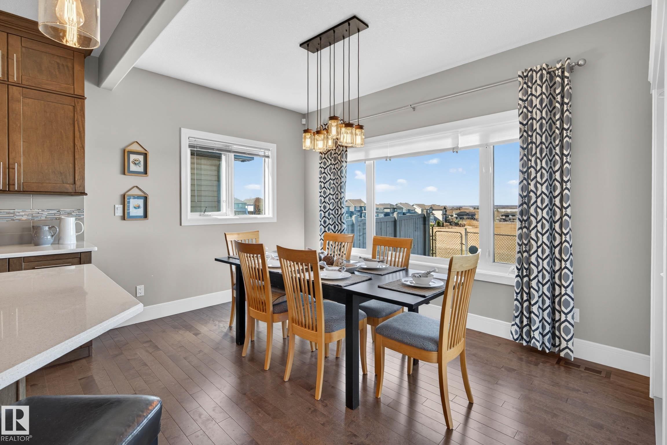 Dining area featuring rich wood-finish flooring, a contemporary linear chandelier, expansive windows, light gray wall tones, and a kitchen counter with a light-toned surface - 2086 Redtail Common, Edmonton, AB - Indoor Photo Showing Dining Room