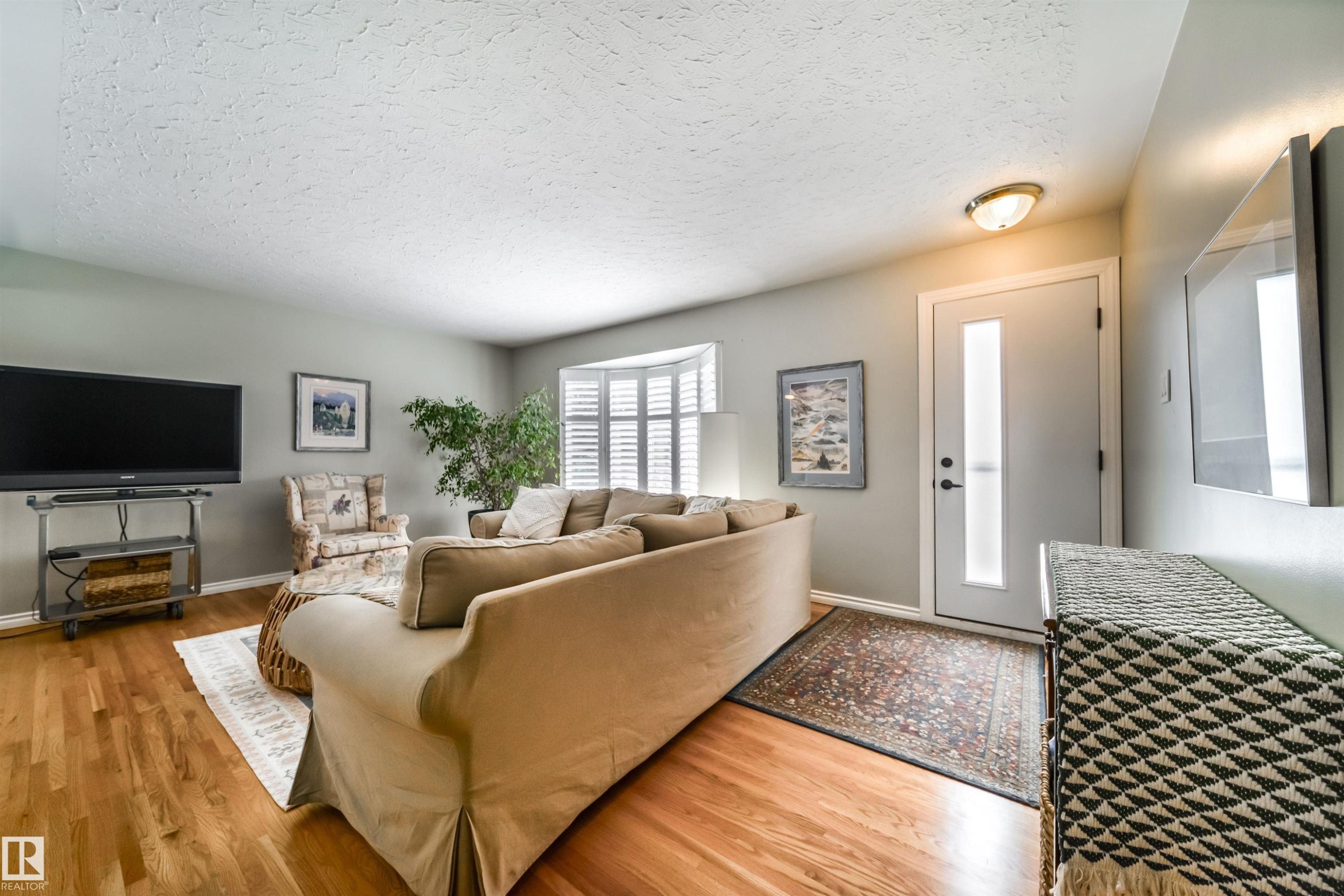 Living area featuring wood-finish flooring, a bay window with plantation shutters, a white entry door with frosted glass insert, and a flush-mount ceiling light fixture - 10231 46 Street, Edmonton, AB - Indoor Photo Showing Living Room