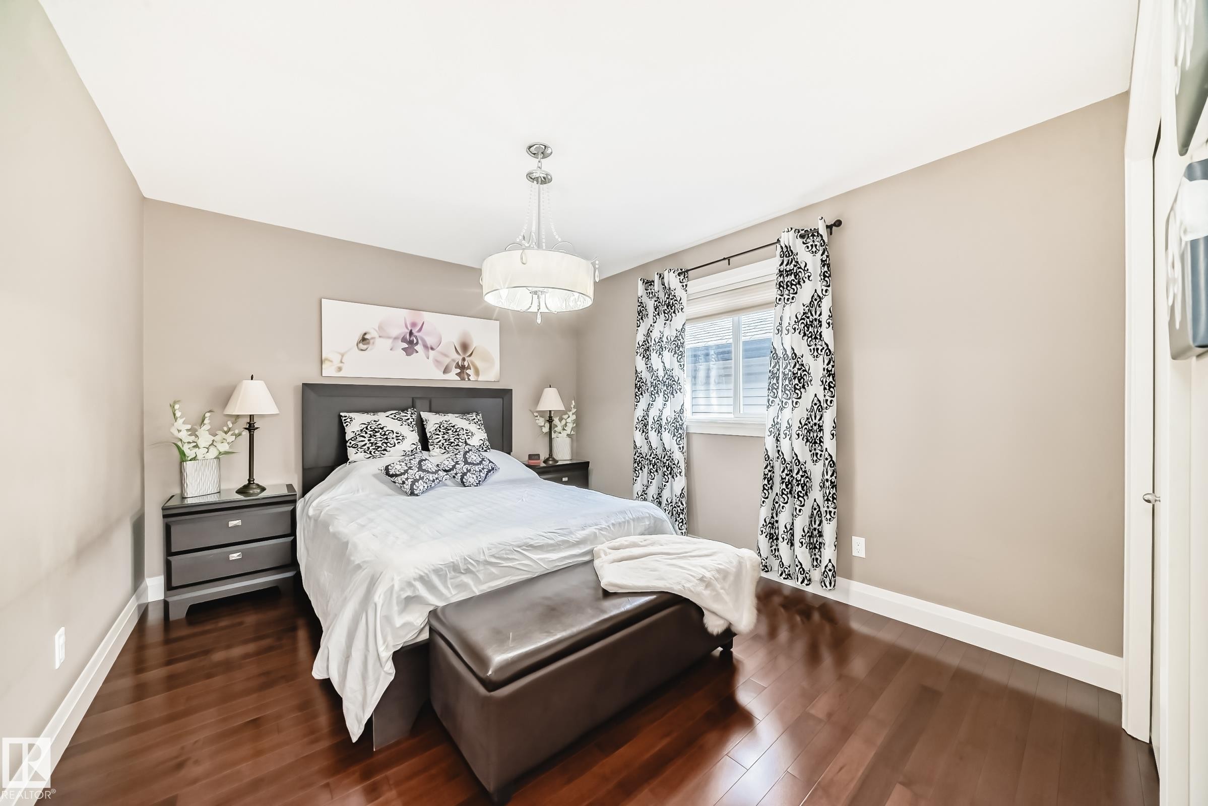 This bedroom features hardwood flooring, a window with white blinds and patterned curtains, and a chandelier-style light fixture - 3220 151 Avenue, Edmonton, AB - Indoor Photo Showing Bedroom