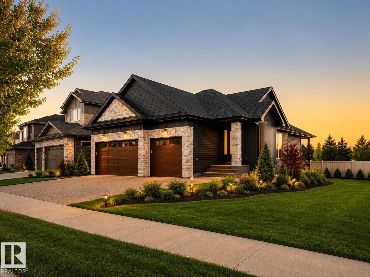 Modern Craftsman-style exterior featuring a dark facade with stone accents, twin wood-panel garage doors, a covered porch with stone columns, and manicured landscaping with integrated lighting - 149 Eastgate Way, St. Albert, AB - Outdoor With Facade