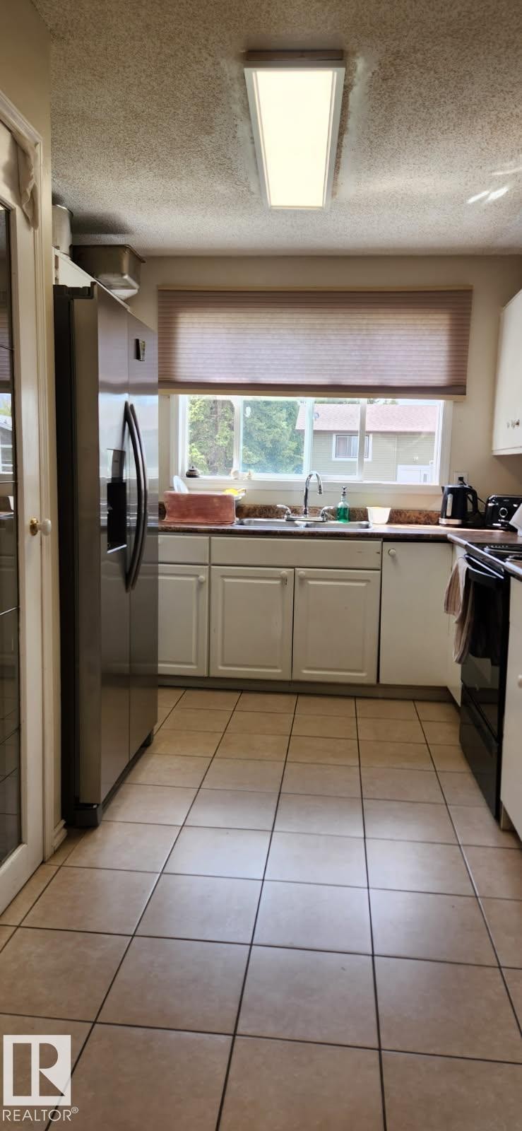Galley kitchen featuring light tile flooring, white cabinetry, a stainless steel refrigerator, and a window above the sink - 14411 31 Street, Edmonton, AB - Indoor