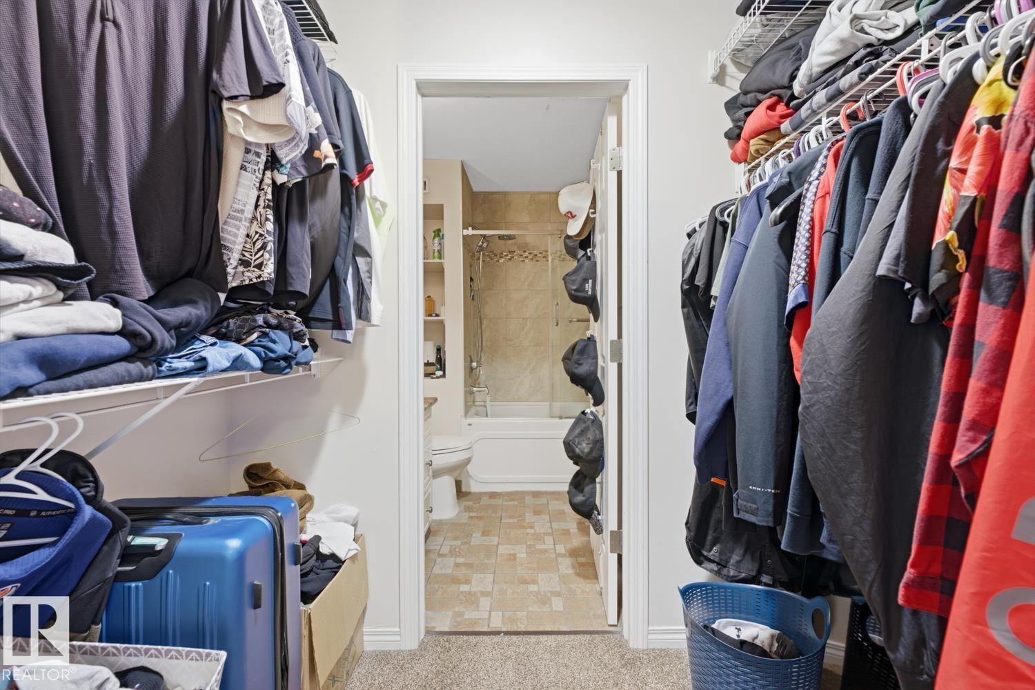 Walk-in closet with carpet flooring and direct access to a full bathroom featuring tile flooring, a built-in bathtub, and a tiled shower surround - #115 10105 95 St Nw, Edmonton, AB - Indoor With Storage
