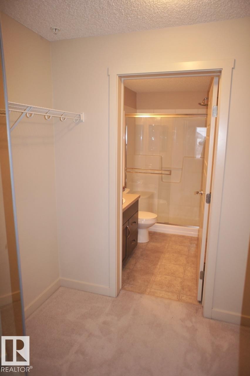 Carpeted dressing area with built-in wire shelving and direct access to a full bathroom featuring a vanity with storage, a toilet, a shower with a sliding glass door, and tiled flooring - 326 6076 Schonsee Way, Edmonton, AB - Indoor Photo Showing Bathroom