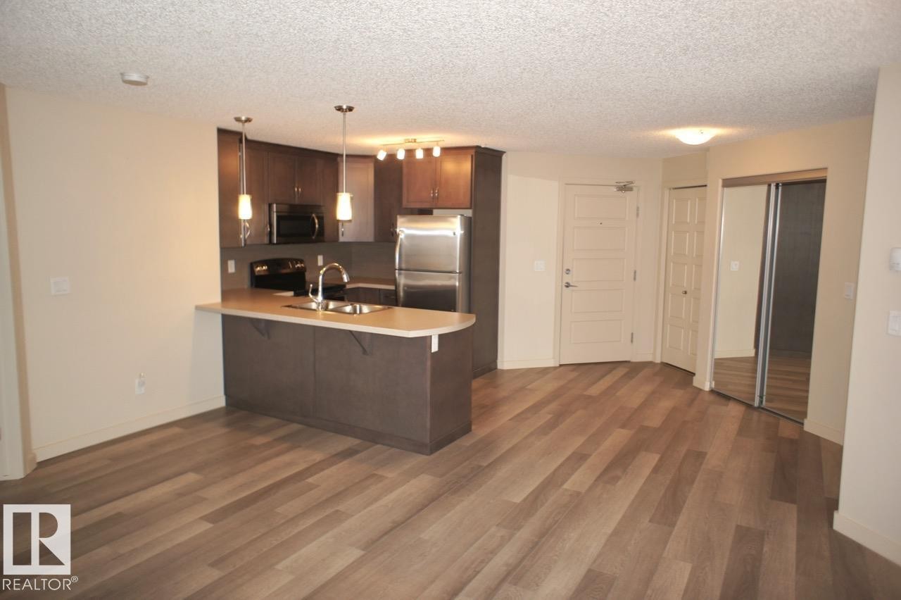 Open-concept living space with wood-finish flooring, a kitchen island with pendant lighting, stainless steel appliances, and mirrored closet doors - 326 6076 Schonsee Way, Edmonton, AB - Indoor Photo Showing Kitchen