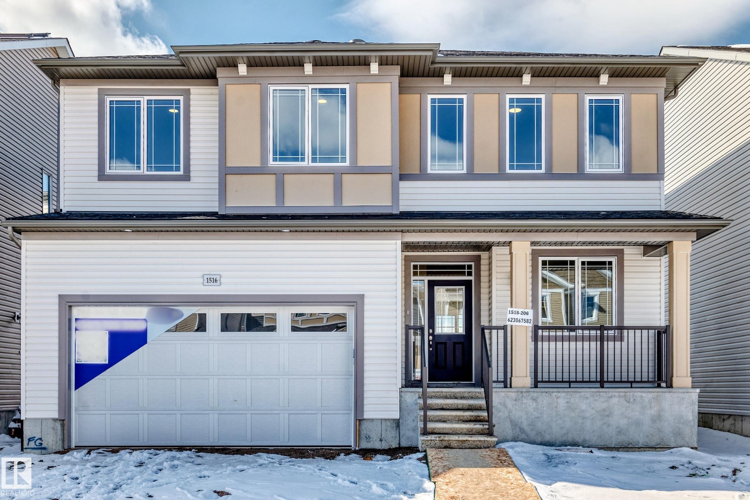 The property features a two-story exterior with light-colored siding, a front entry with a dark door, and a covered porch with dark railings - 1516 206 Street Nw, Edmonton, AB - Outdoor