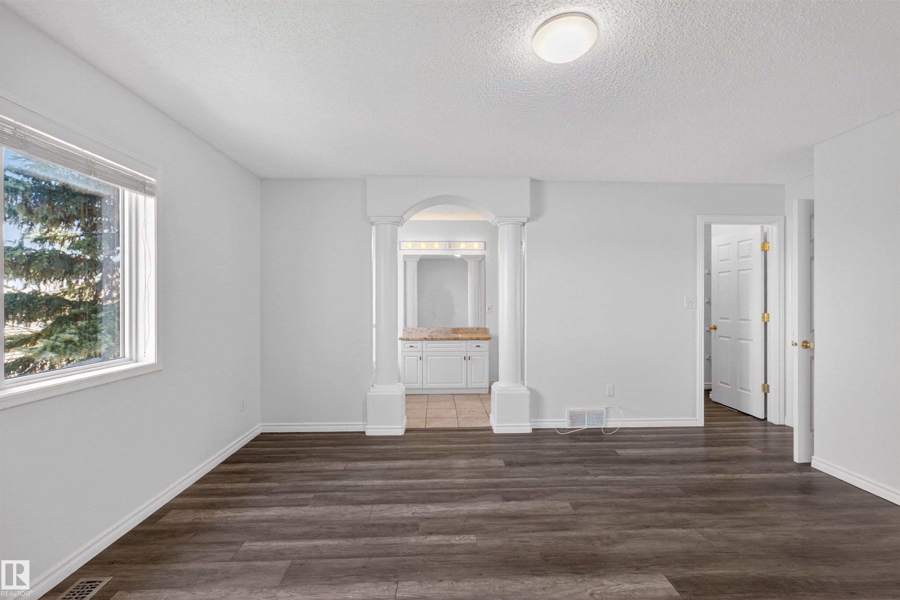 Spacious room featuring wood-finish flooring, a large window, and an arched entry with decorative columns leading to a vanity area - 6 1237 Carter Crest Road, Edmonton, AB