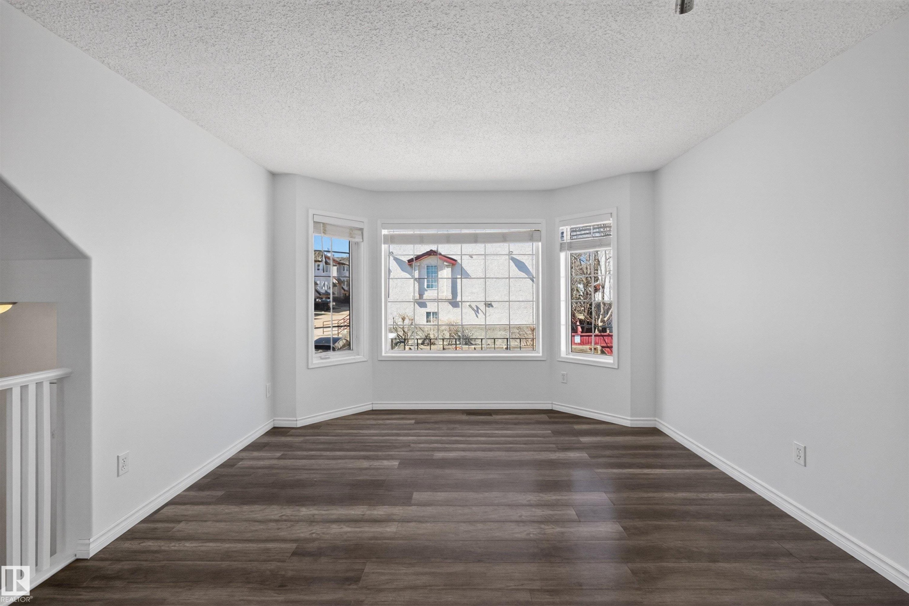 Bright interior room featuring a bay window, wood-finish flooring, white baseboards, a textured ceiling, and a white railing - 6 1237 Carter Crest Road, Edmonton, AB