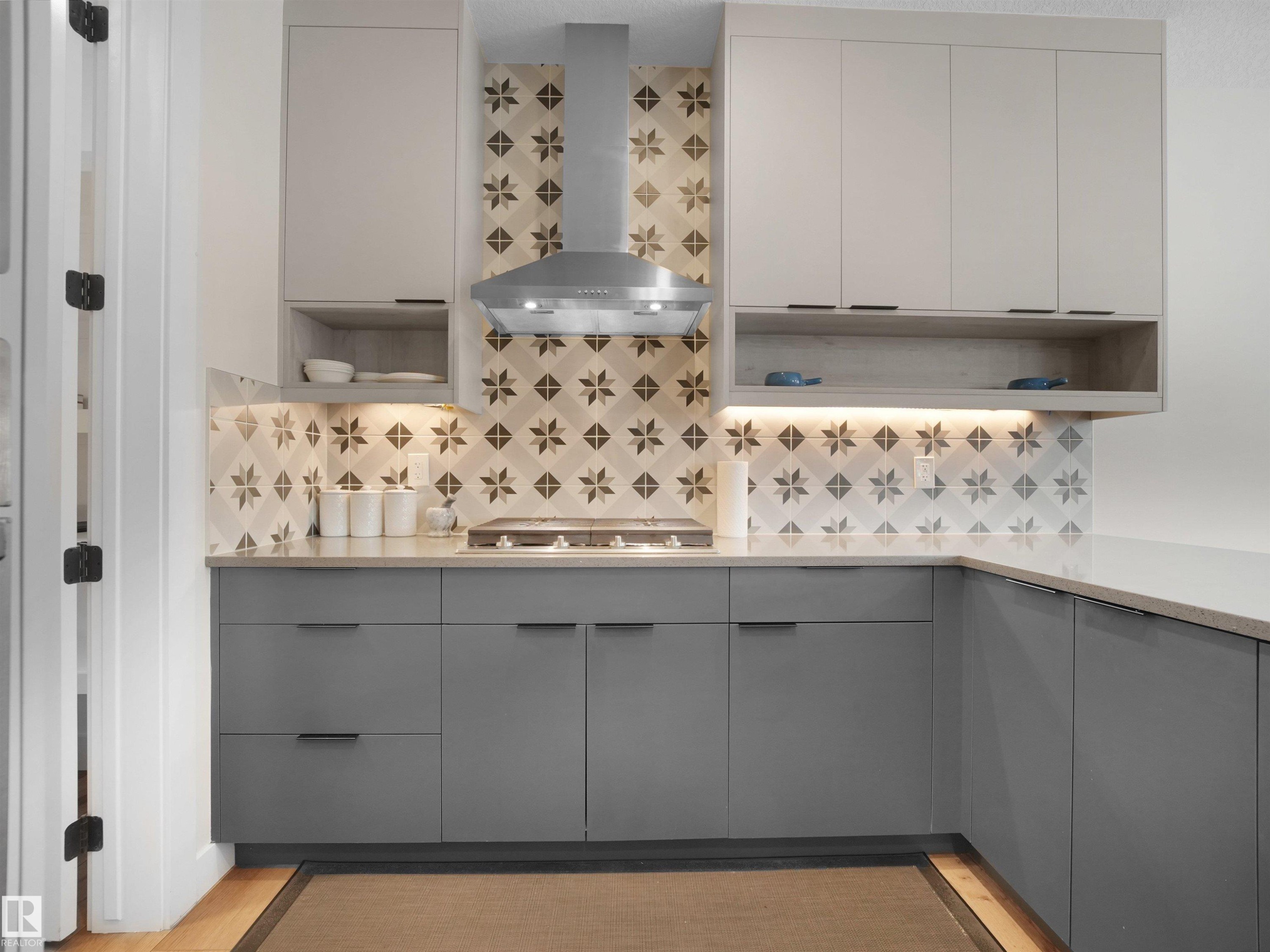 Modern kitchen featuring gray base cabinetry, light upper cabinetry with open shelving, a patterned tile backsplash, stainless steel range hood, and light-toned countertops - 20827 130 Avenue Nw, Edmonton, AB - Indoor