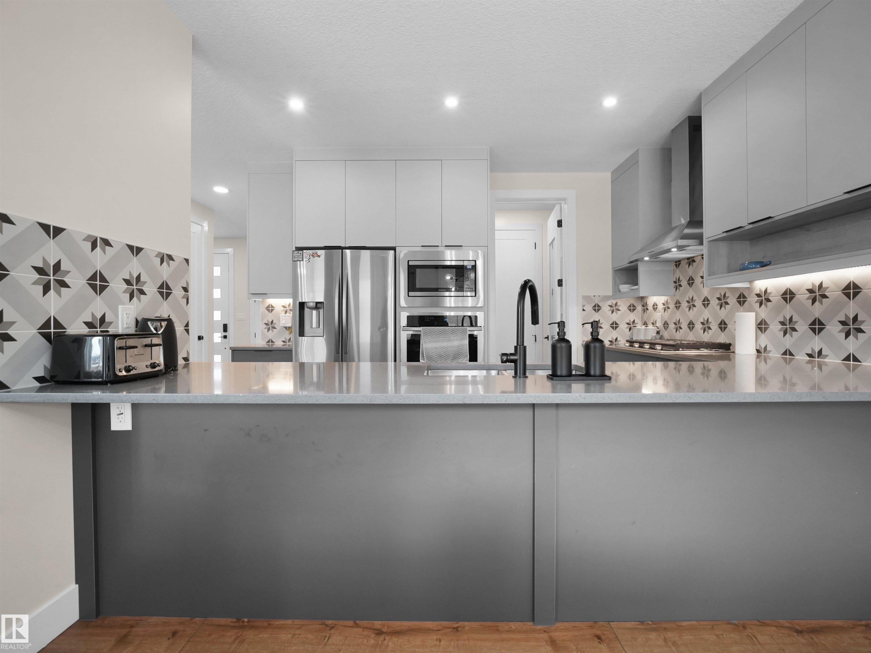 Kitchen featuring recessed lighting, a built-in stainless steel microwave, a wall-mounted range hood, a patterned tile backsplash, and wood-finish flooring - 20827 130 Avenue Nw, Edmonton, AB - Indoor Photo Showing Kitchen With Upgraded Kitchen