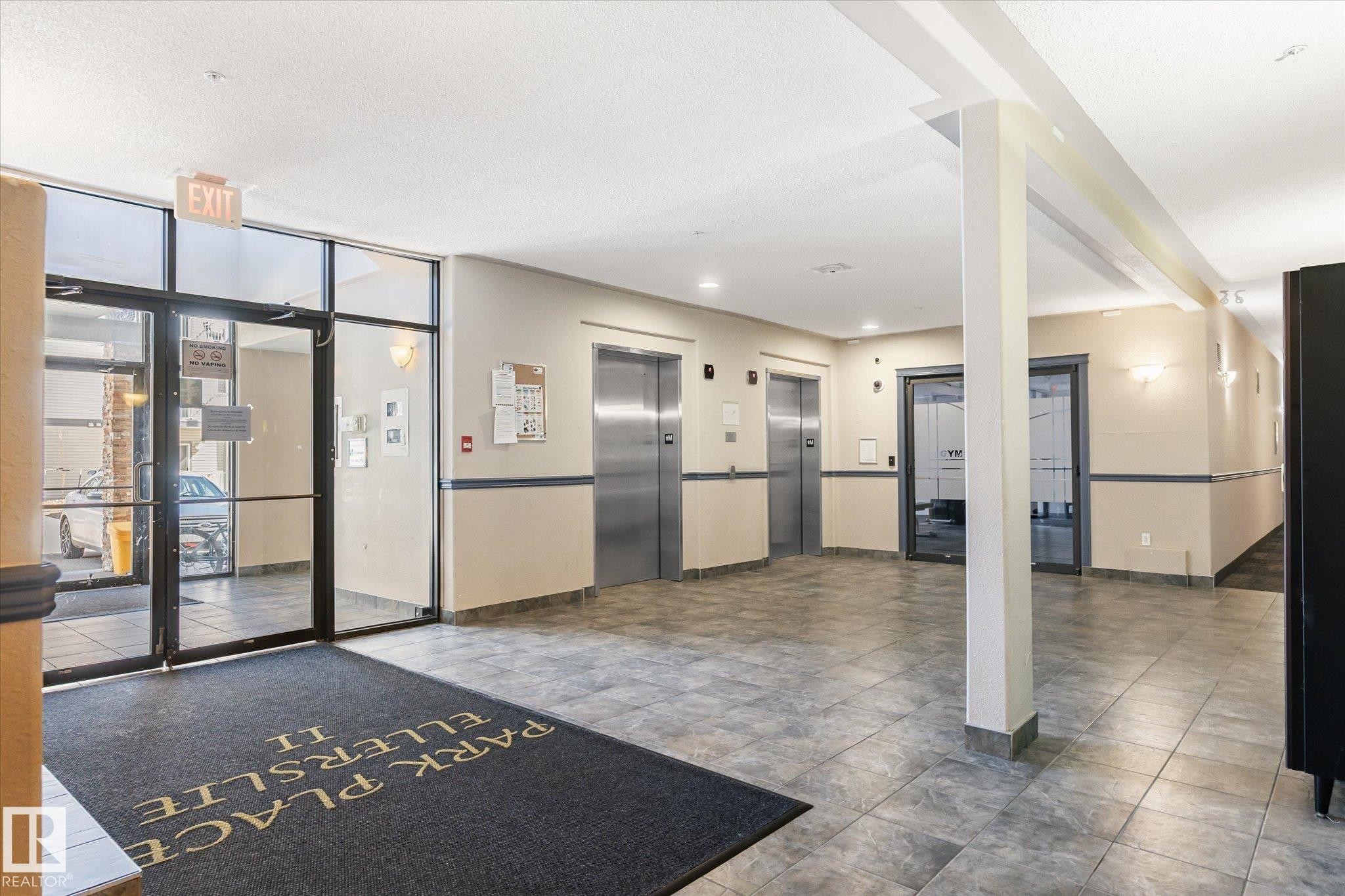 Building lobby with multiple stainless steel elevators, tiled flooring, neutral-toned walls, and glass entry doors - 203 155 Edwards Drive, Edmonton, AB - Indoor Photo Showing Other Room