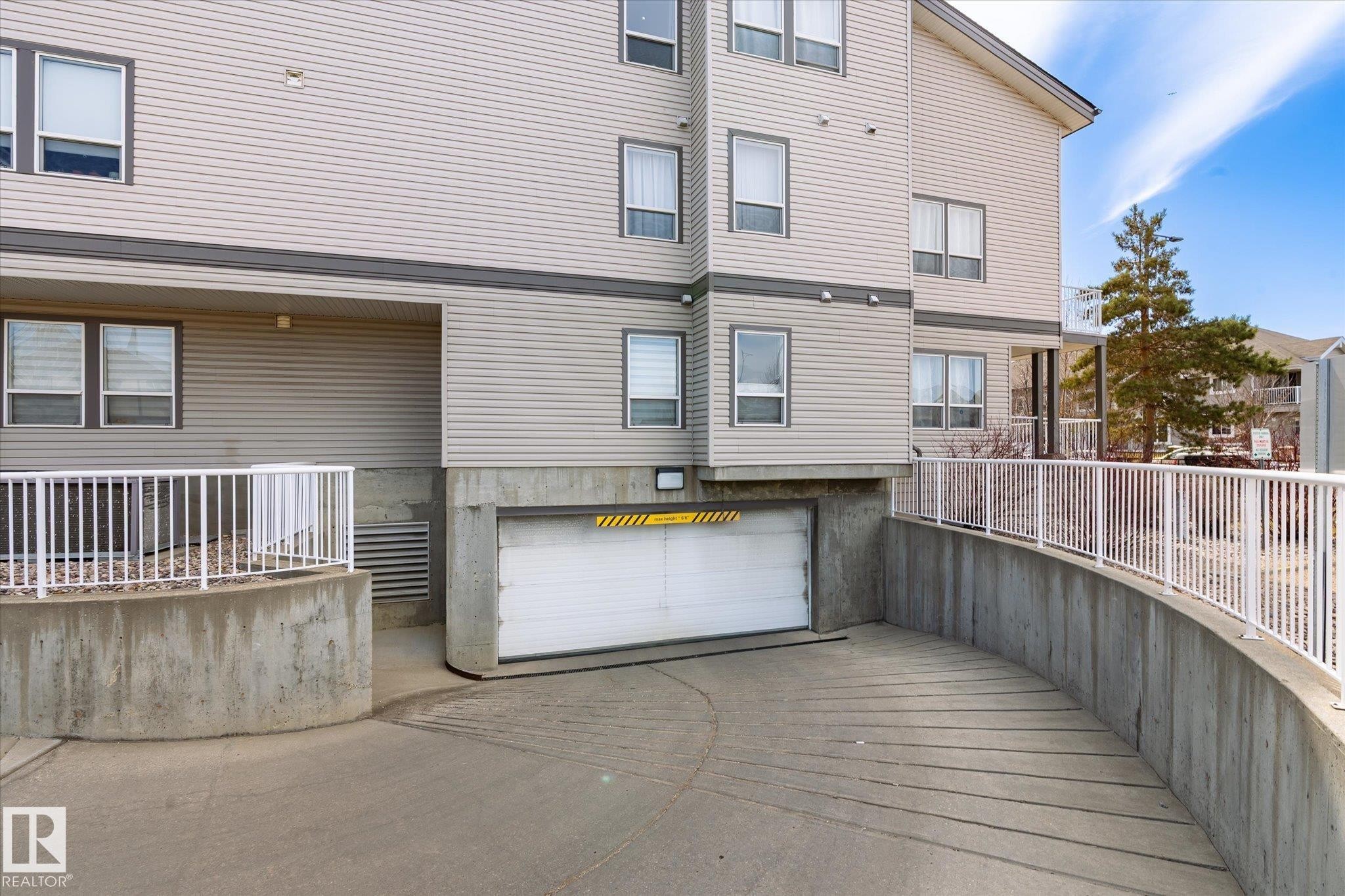 Building exterior featuring light-colored siding, multiple windows with blinds, an attached garage with a white door, a concrete access ramp, and white metal railings - 203 155 Edwards Drive, Edmonton, AB - Outdoor With Exterior