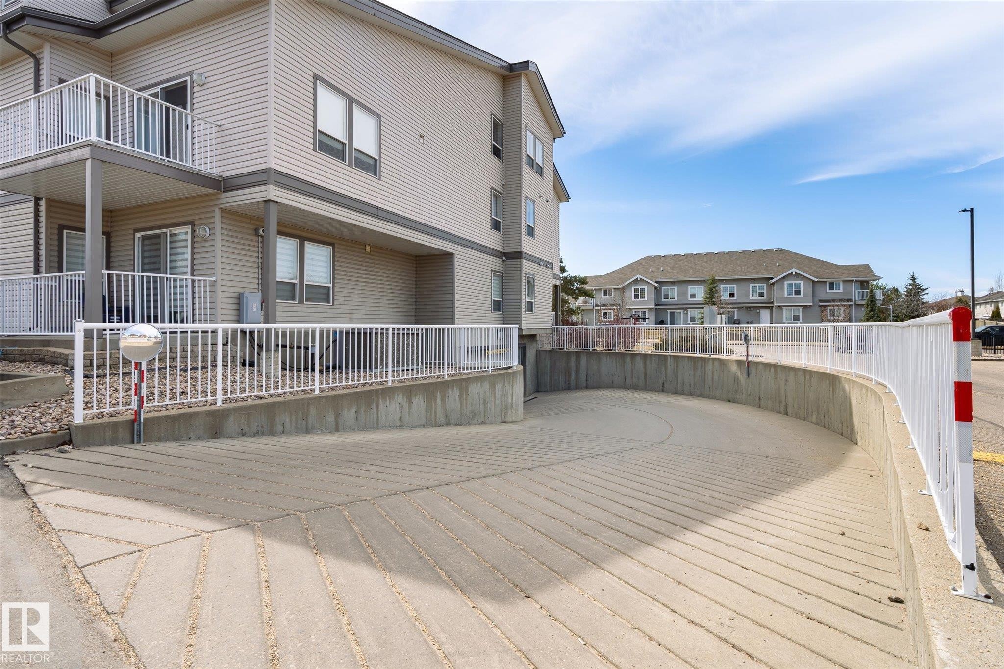 Curved concrete ramp access to underground parking, featuring a white metal railing and a convex safety mirror - 203 155 Edwards Drive, Edmonton, AB - Outdoor With Balcony