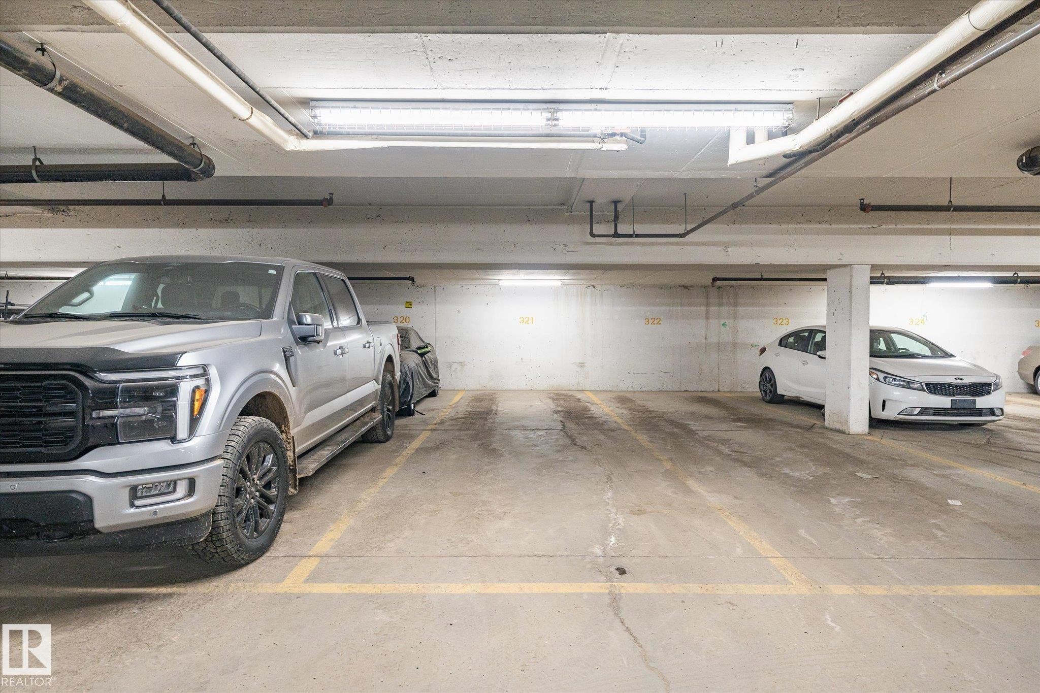 Underground parking space with concrete flooring, overhead fluorescent lighting, and exposed ceiling pipes - 203 155 Edwards Drive, Edmonton, AB - Indoor Photo Showing Garage