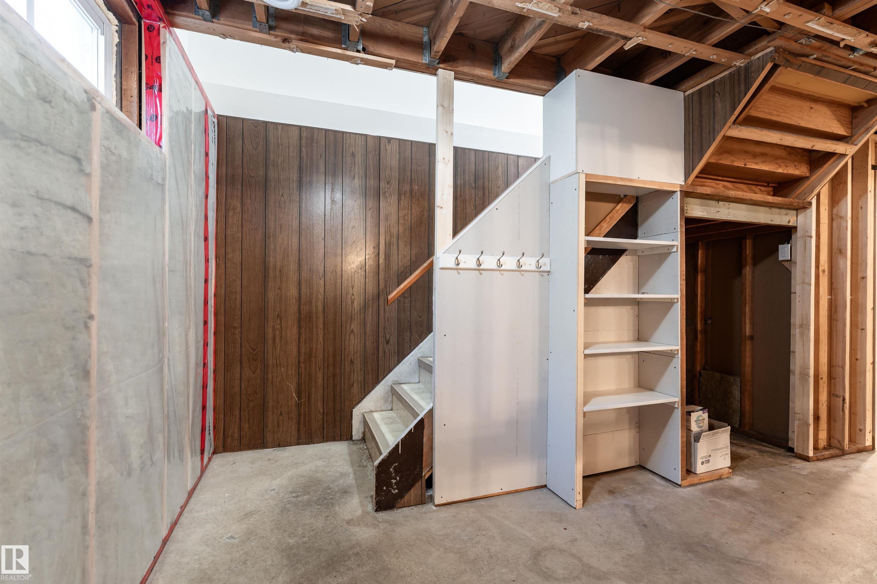Unfinished basement area featuring exposed ceiling joists, concrete flooring, wood paneling, an unfinished staircase with a wood handrail, and built-in shelving - 13331 64 Street Nw, Edmonton, AB - Indoor Photo Showing Basement