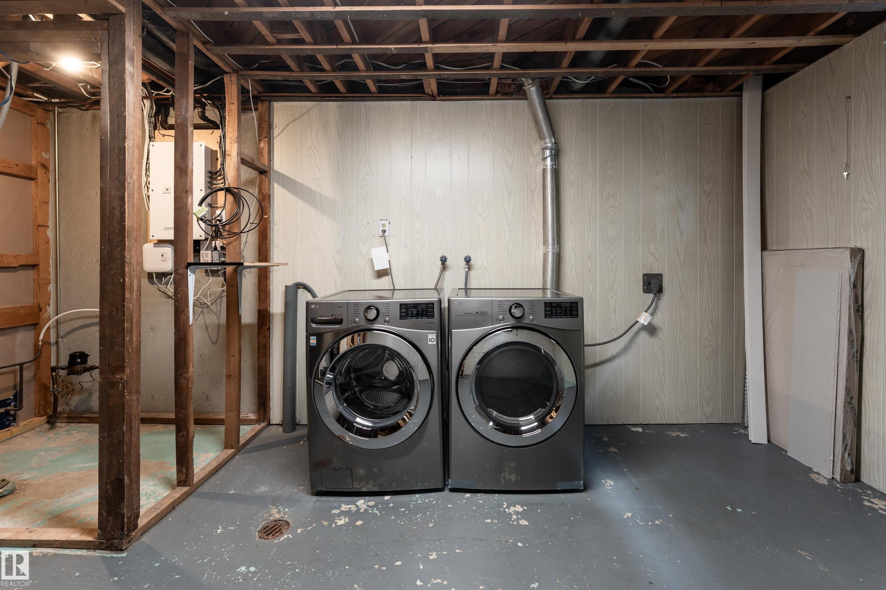 Laundry area featuring a side-by-side washer and dryer, painted concrete flooring, wall paneling, exposed ceiling joists, and visible utility connections - 13331 64 Street Nw, Edmonton, AB - Indoor Photo Showing Laundry Room