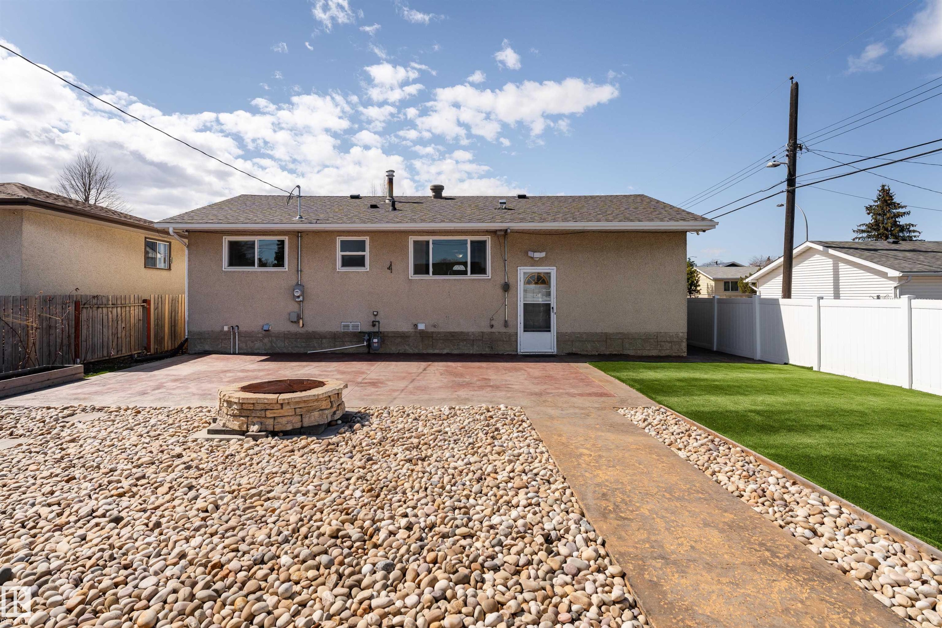 Expansive rear yard featuring a stone fire pit, stamped concrete patio, artificial turf, and a white vinyl fence - 13331 64 Street Nw, Edmonton, AB - Outdoor