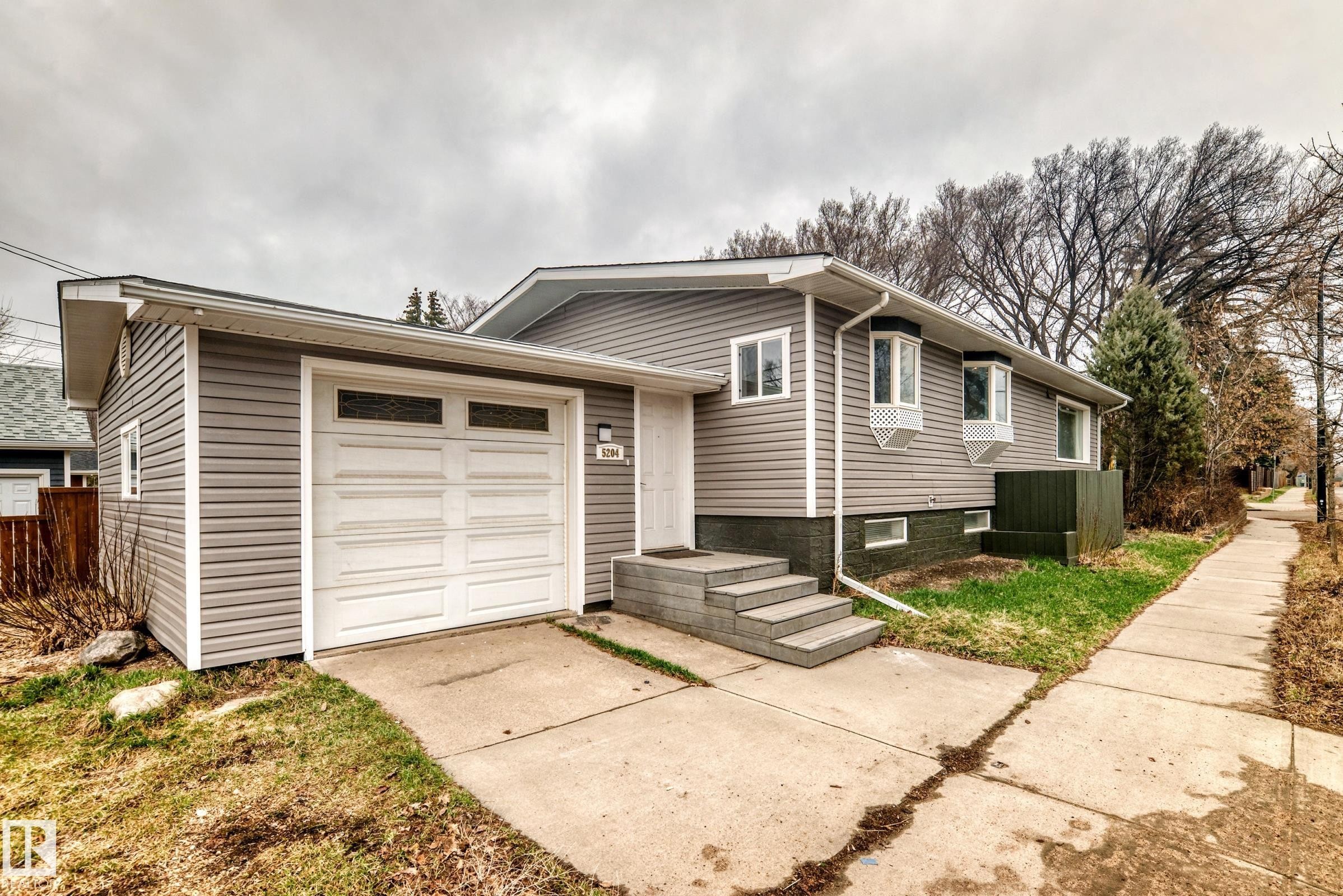 Split-level residence featuring grey horizontal siding, a white garage door with upper window inserts, and concrete steps leading to the main entrance - 5204 112 Avenue Nw, Edmonton, AB - Outdoor