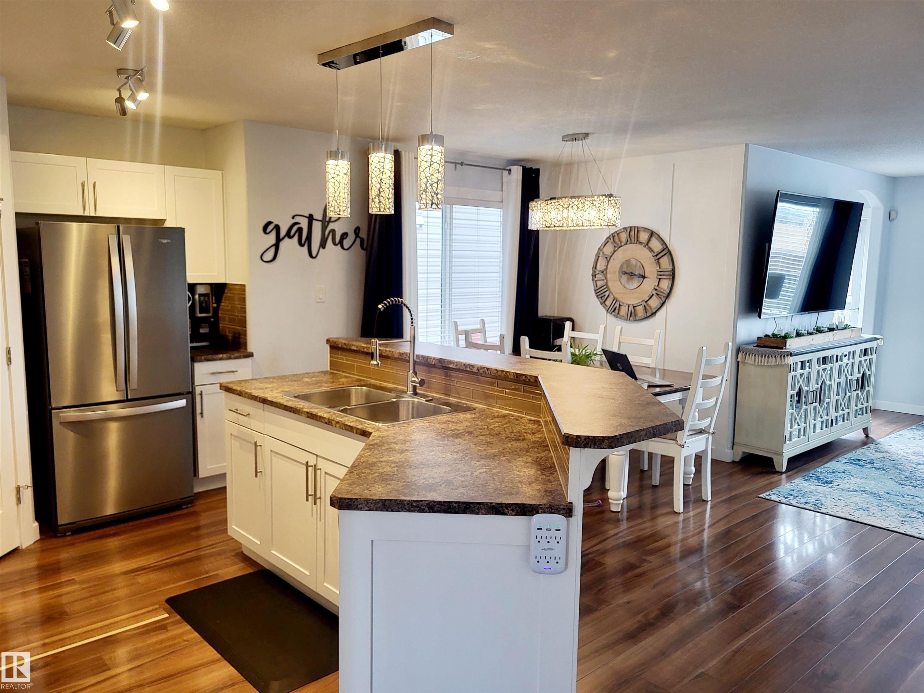 Open-concept kitchen featuring an island with a double basin sink, white cabinetry, a stainless steel refrigerator, and wood-finish flooring - 144 Kirpatrick Way, Leduc, AB - Indoor Photo Showing Kitchen With Double Sink