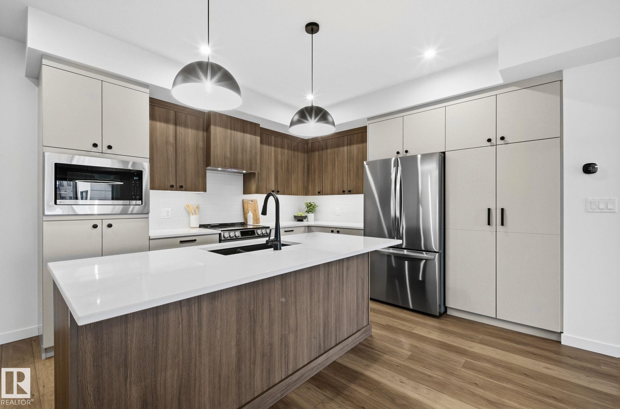 Modern kitchen featuring a large central island with a white countertop, two-tone cabinetry, stainless steel appliances, and wood-finish flooring - 202 7459 May Common, Edmonton, AB - Indoor Photo Showing Kitchen With Upgraded Kitchen