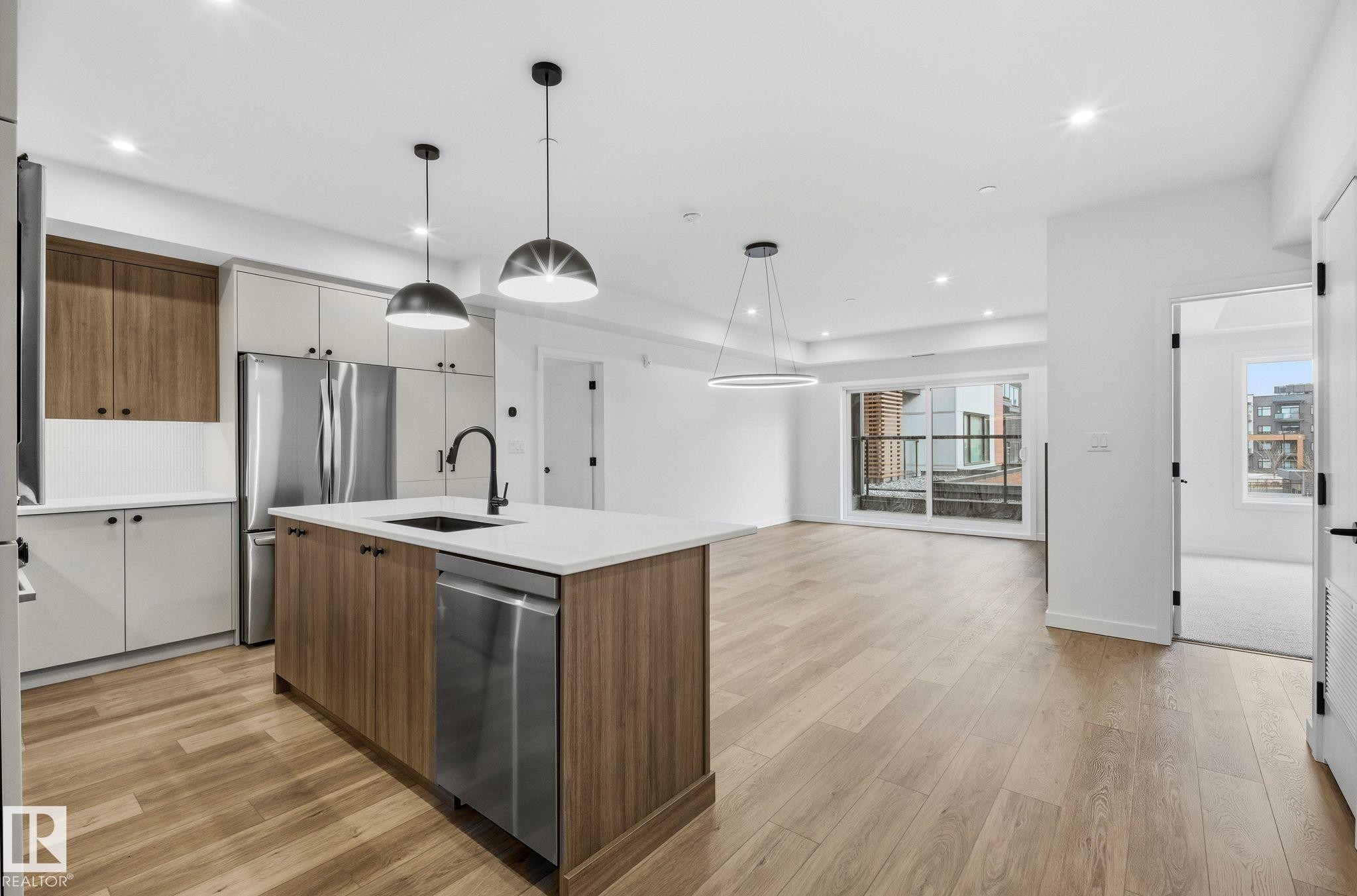 Modern kitchen with a central island featuring a stainless steel sink, integrated dishwasher, and pendant lighting - 202 7459 May Common, Edmonton, AB - Indoor Photo Showing Kitchen With Upgraded Kitchen