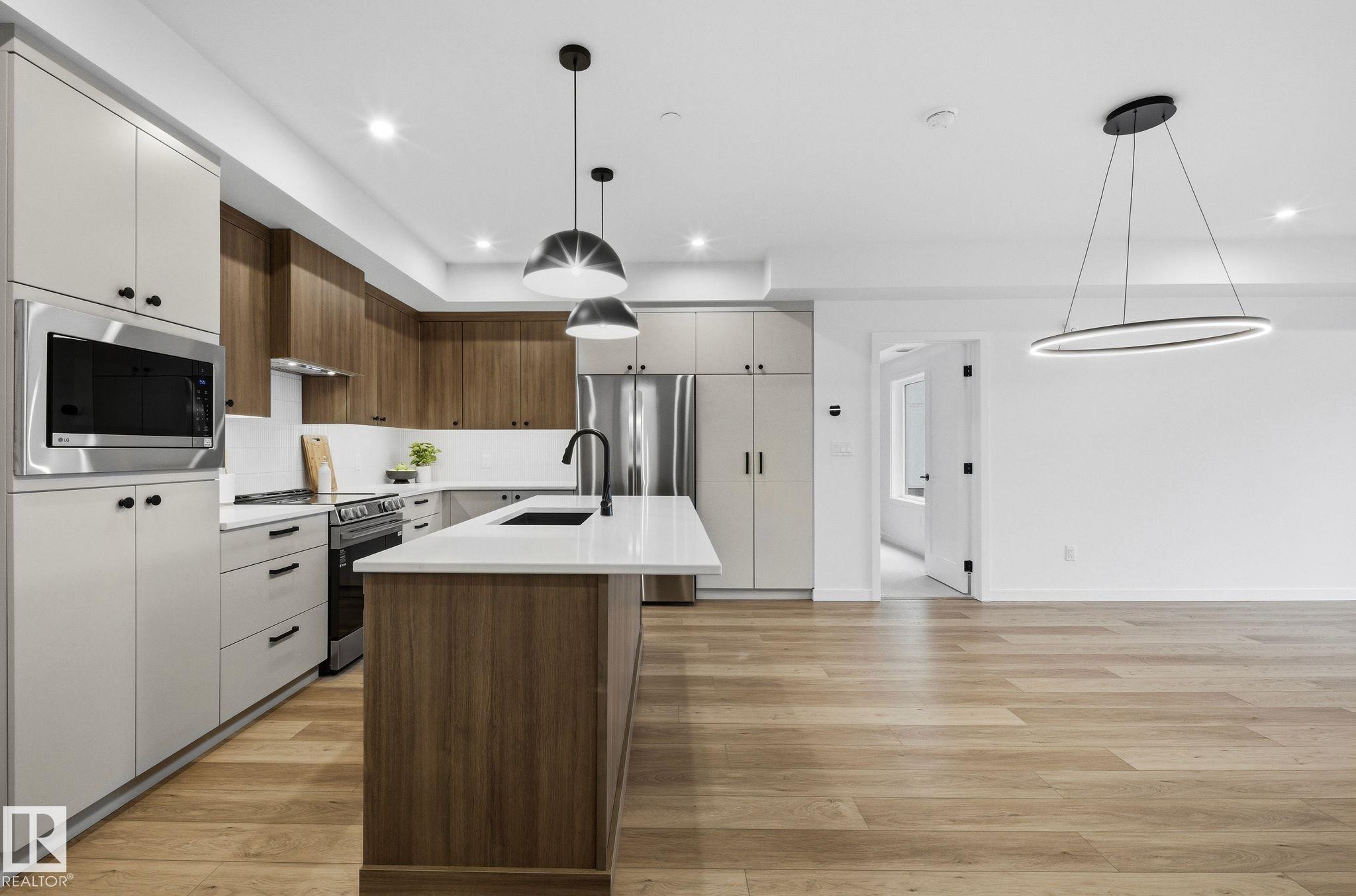 Modern kitchen featuring two-tone cabinetry, stainless steel appliances, a central island with a sink, and pendant lighting - 202 7459 May Common, Edmonton, AB - Indoor Photo Showing Kitchen With Upgraded Kitchen