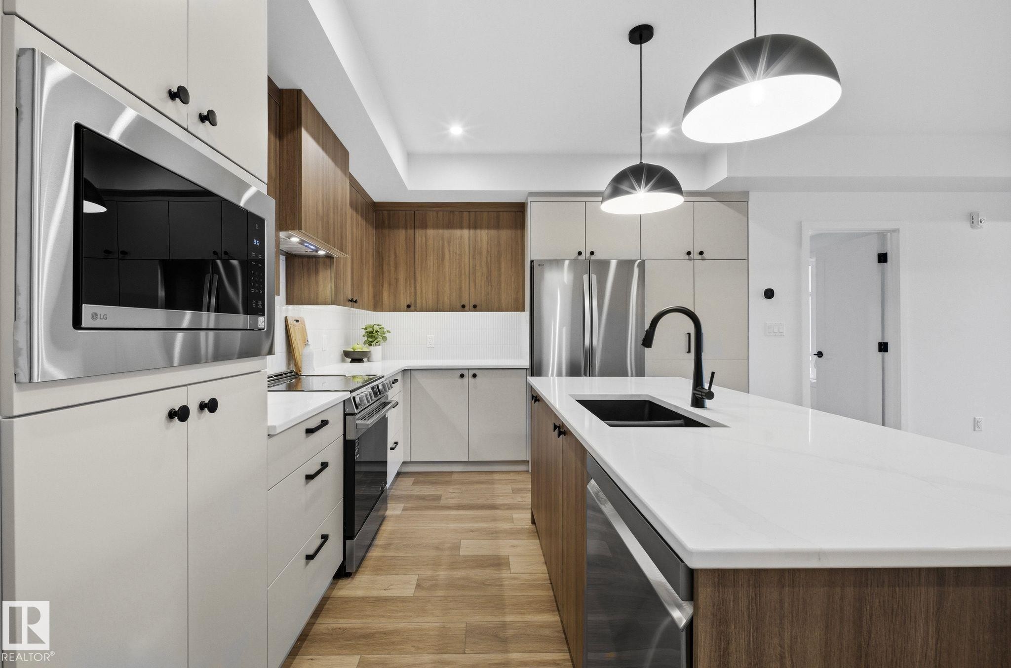 Modern kitchen featuring an extended island with a black sink and matte black faucet, stainless steel appliances, wood-finish flooring, recessed lighting, and elegant pendant fixtures - 202 7459 May Common, Edmonton, AB - Indoor Photo Showing Kitchen With Double Sink With Upgraded Kitchen
