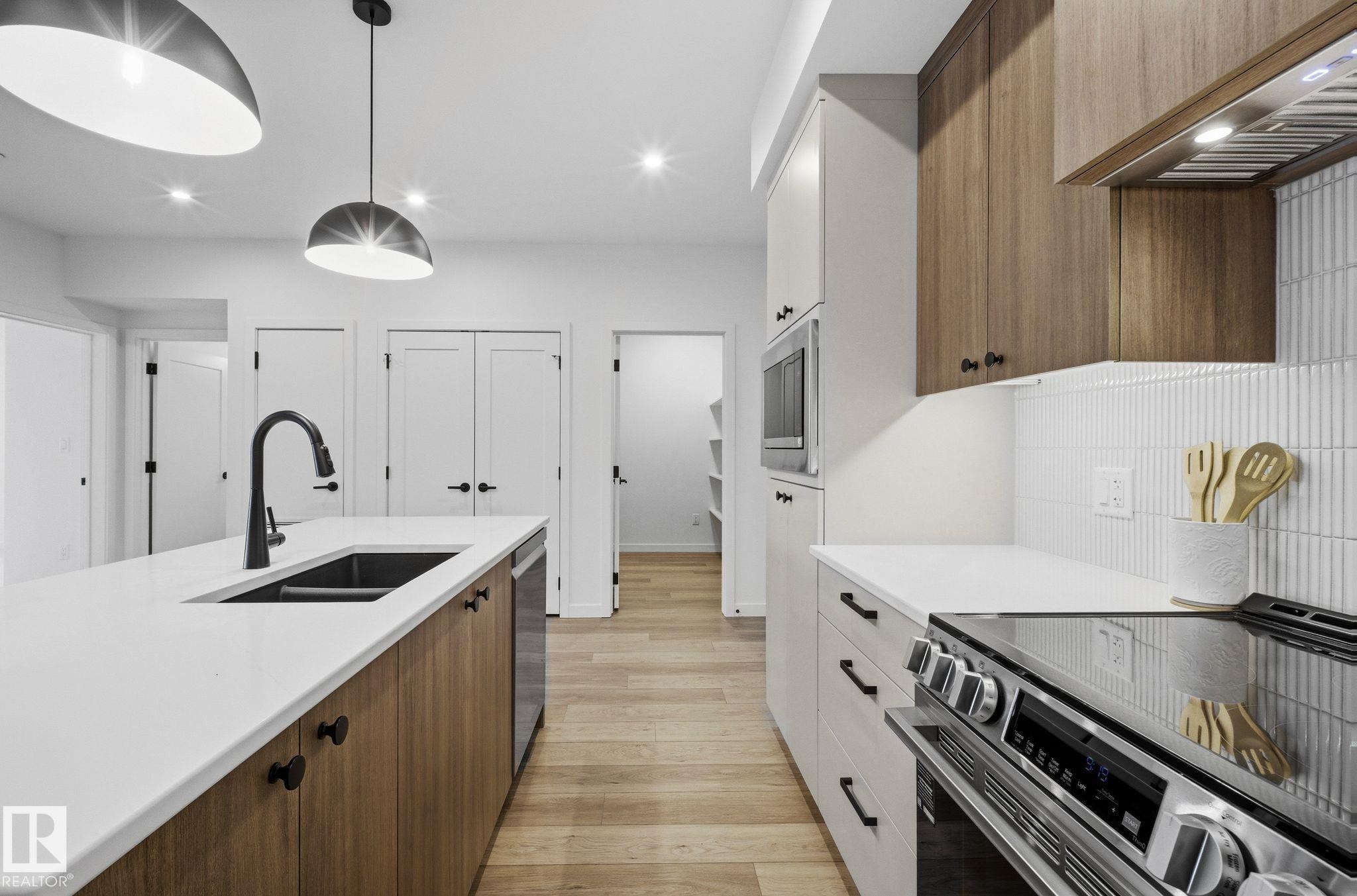 Modern kitchen featuring a large island with an undermount sink and matte black faucet, wood-finish base cabinets, and white countertops - 202 7459 May Common, Edmonton, AB - Indoor Photo Showing Kitchen With Double Sink With Upgraded Kitchen