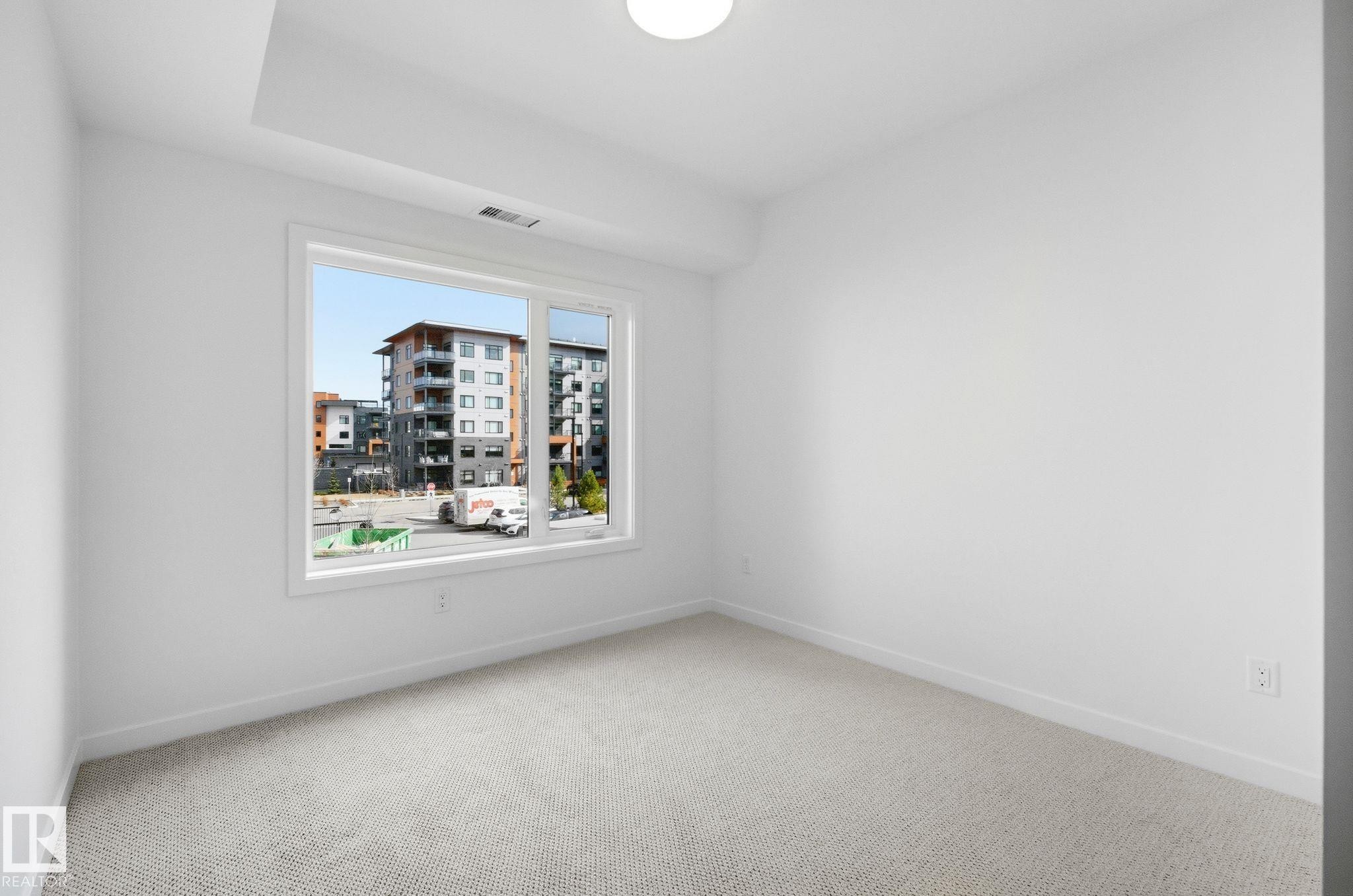 Bright interior space featuring a large window, light-colored carpeting, white walls, and a ceiling-mounted light fixture - 202 7459 May Common, Edmonton, AB - Indoor Photo Showing Other Room