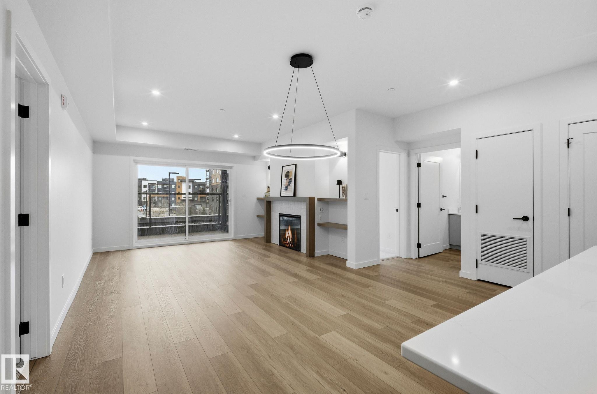 Open-concept living area featuring light wood-finish flooring, a modern fireplace with integrated shelving, and recessed lighting - 202 7459 May Common, Edmonton, AB - Indoor Photo Showing Living Room