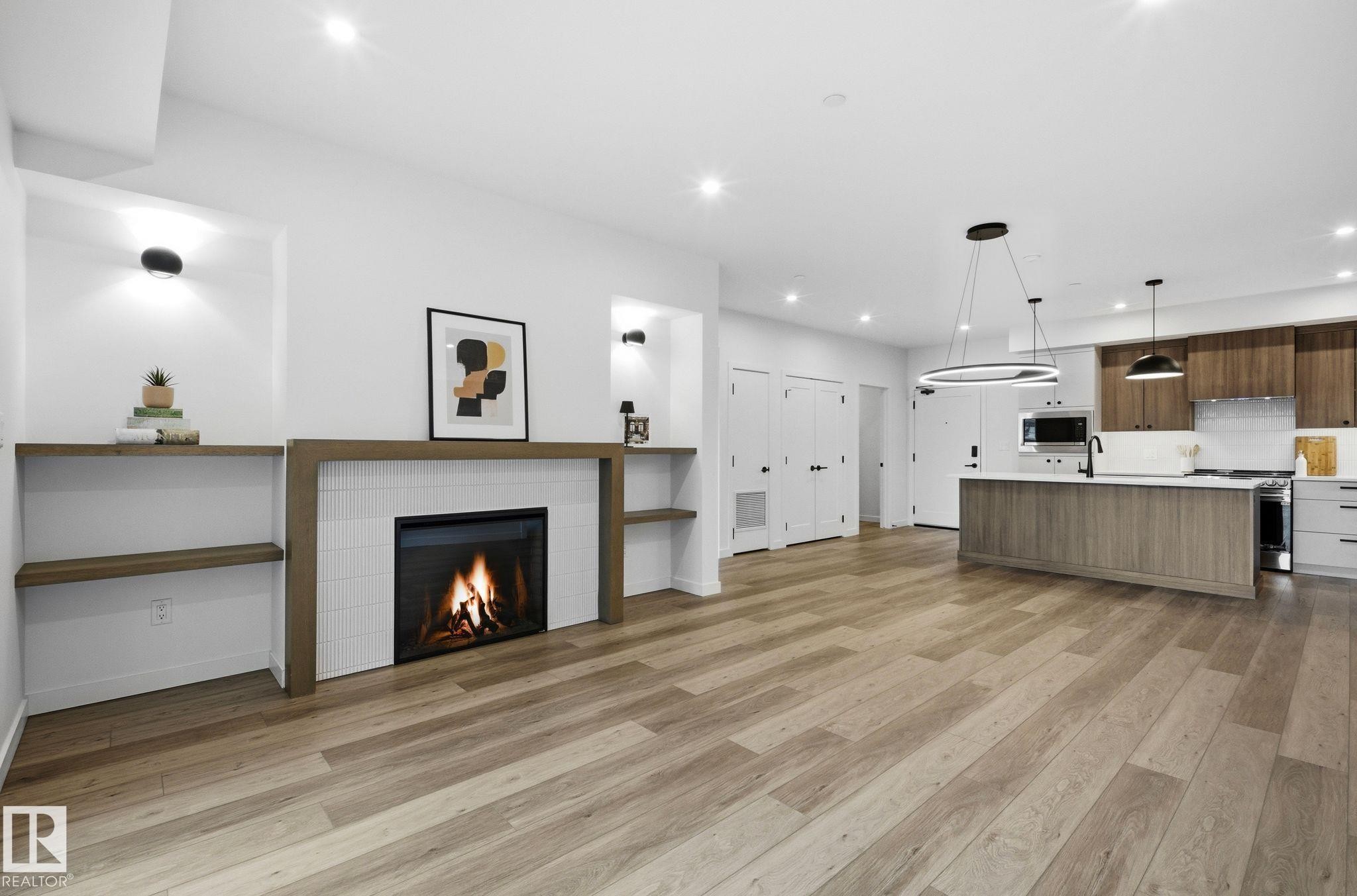 Open-concept living space featuring wood-finish flooring, a modern fireplace with a tiled surround and wood-frame mantel, recessed lighting, and a contemporary kitchen with an island, pendant lighting, and wood cabinetry - 202 7459 May Common, Edmonton, AB - Indoor Photo Showing Other Room With Fireplace