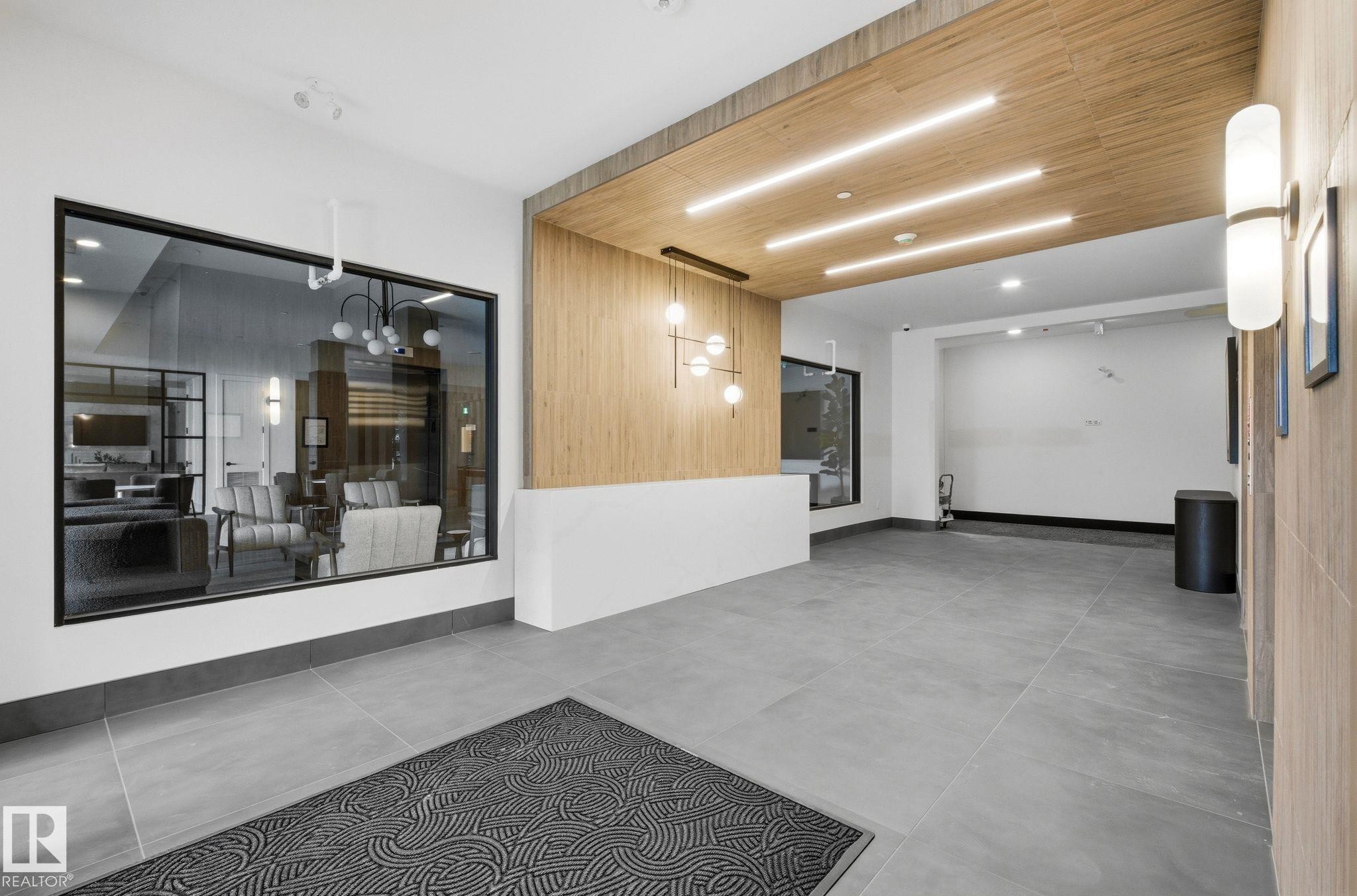 Lobby entry featuring large format grey tile flooring, a wood-paneled ceiling with recessed lighting, and a prominent glass partition - 202 7459 May Common, Edmonton, AB - Indoor Photo Showing Other Room