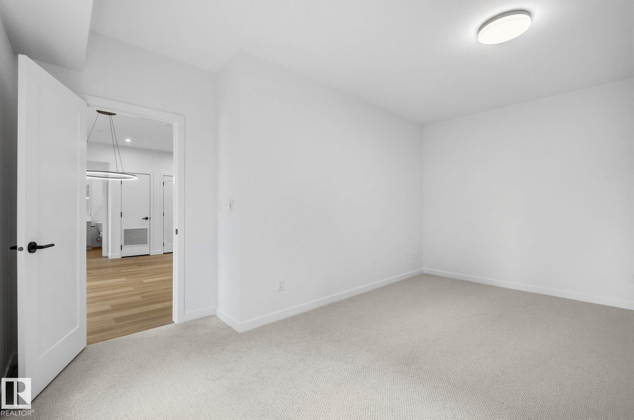 Carpeted room with white walls, a flush-mount ceiling light, and a doorway opening to an area with wood-finish flooring and a contemporary pendant light fixture - 202 7459 May Common, Edmonton, AB - Indoor Photo Showing Other Room