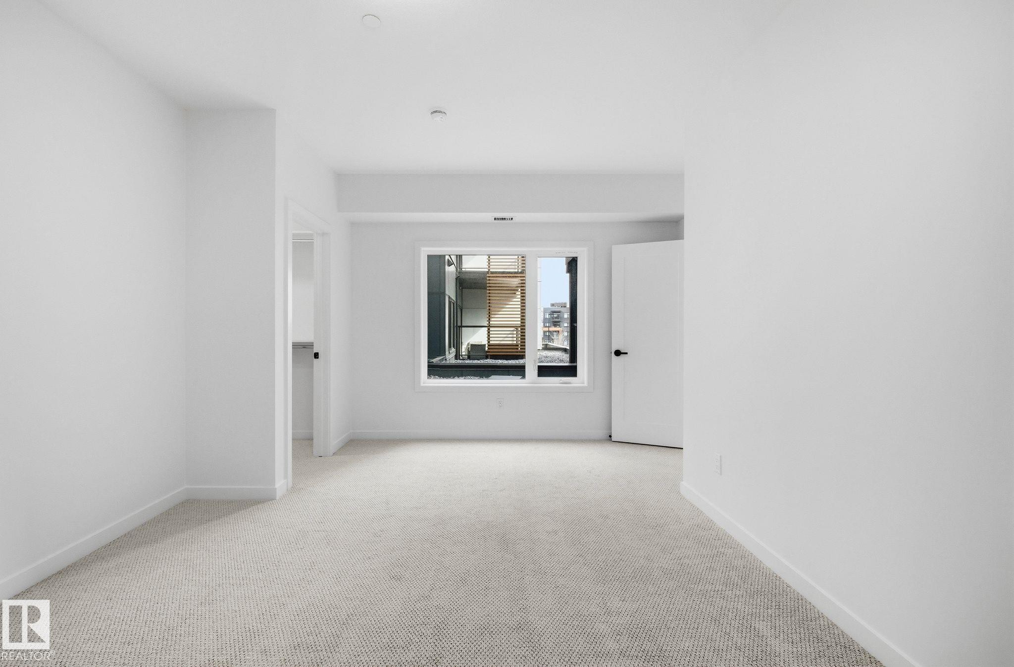 Neutral-toned carpet flooring in a spacious room with a large window, white trim, and a white interior door with black hardware - 202 7459 May Common, Edmonton, AB - Indoor Photo Showing Other Room