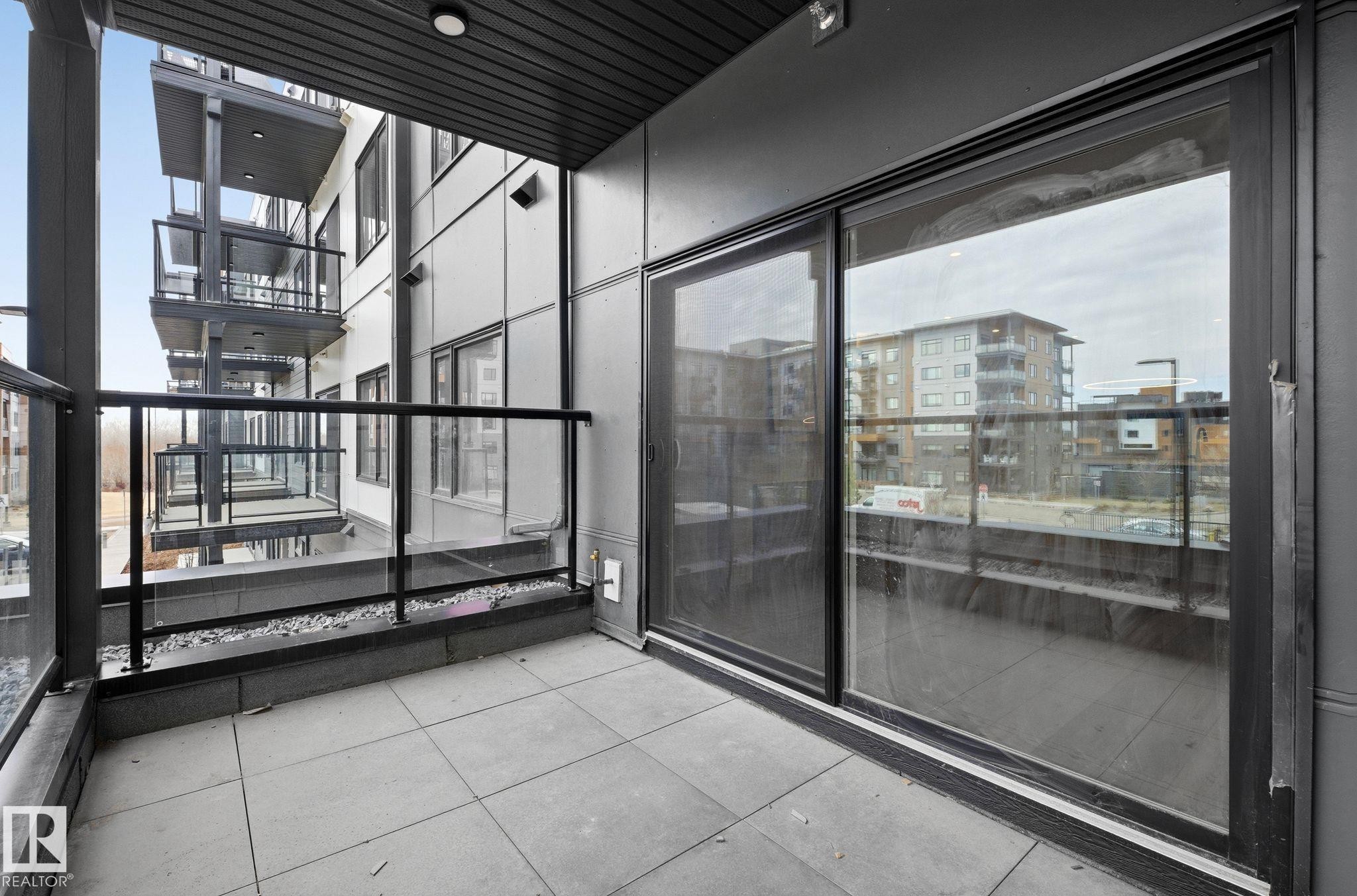 Private balcony featuring square tile flooring and a black metal and glass railing - 202 7459 May Common, Edmonton, AB - Outdoor With Balcony With Exterior