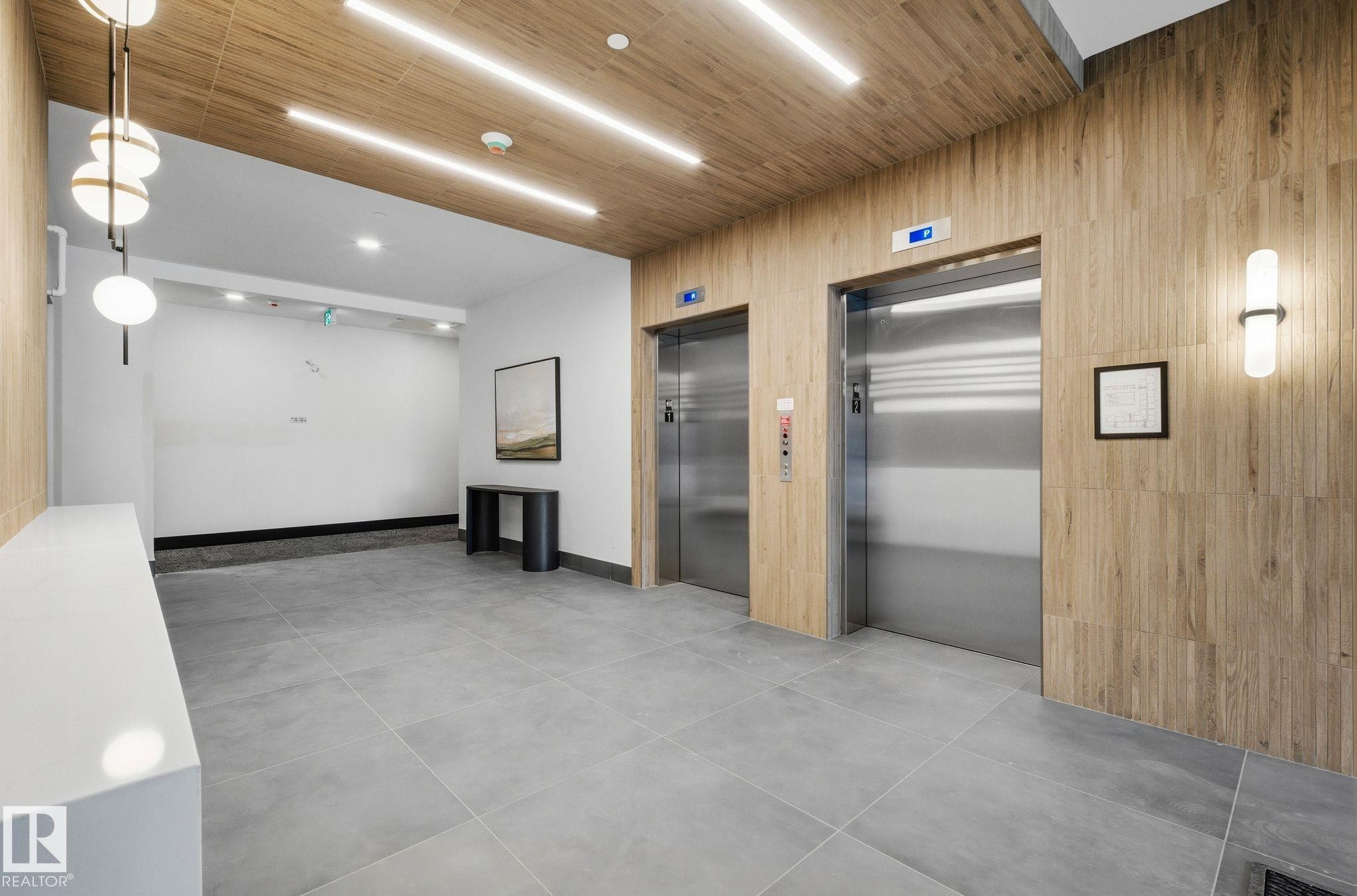 Dual stainless steel elevators set within wood-finish wall panels and a complementary wood-finish ceiling - 202 7459 May Common, Edmonton, AB - Indoor