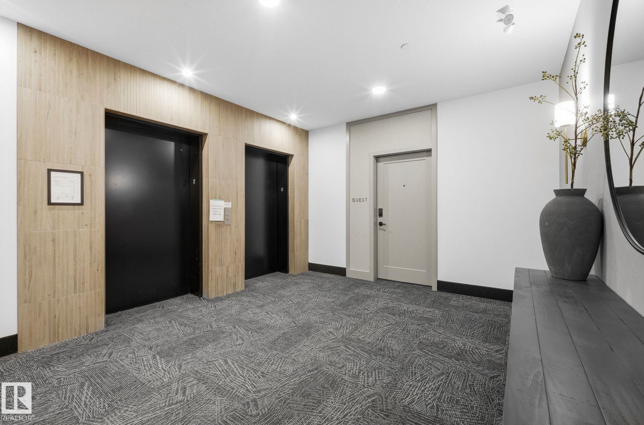 Contemporary hallway featuring two black elevator doors, textured wood-finish wall paneling, and recessed lighting - 202 7459 May Common, Edmonton, AB - Indoor