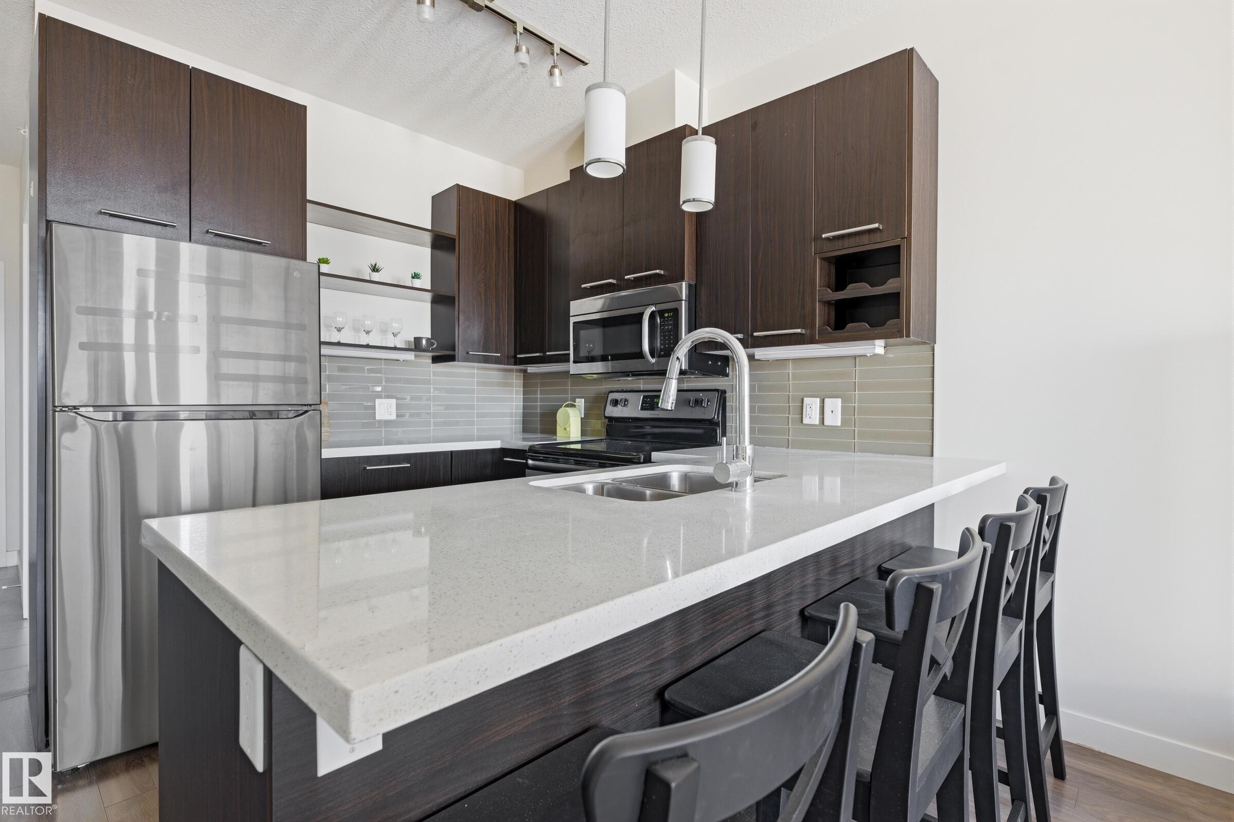 Kitchen featuring a white countertop island with integrated sink, stainless steel appliances, dark wood-finish cabinetry, and a horizontal tile backsplash - 403 10418 81 Avenue, Edmonton, AB - Indoor Photo Showing Kitchen With Double Sink With Upgraded Kitchen
